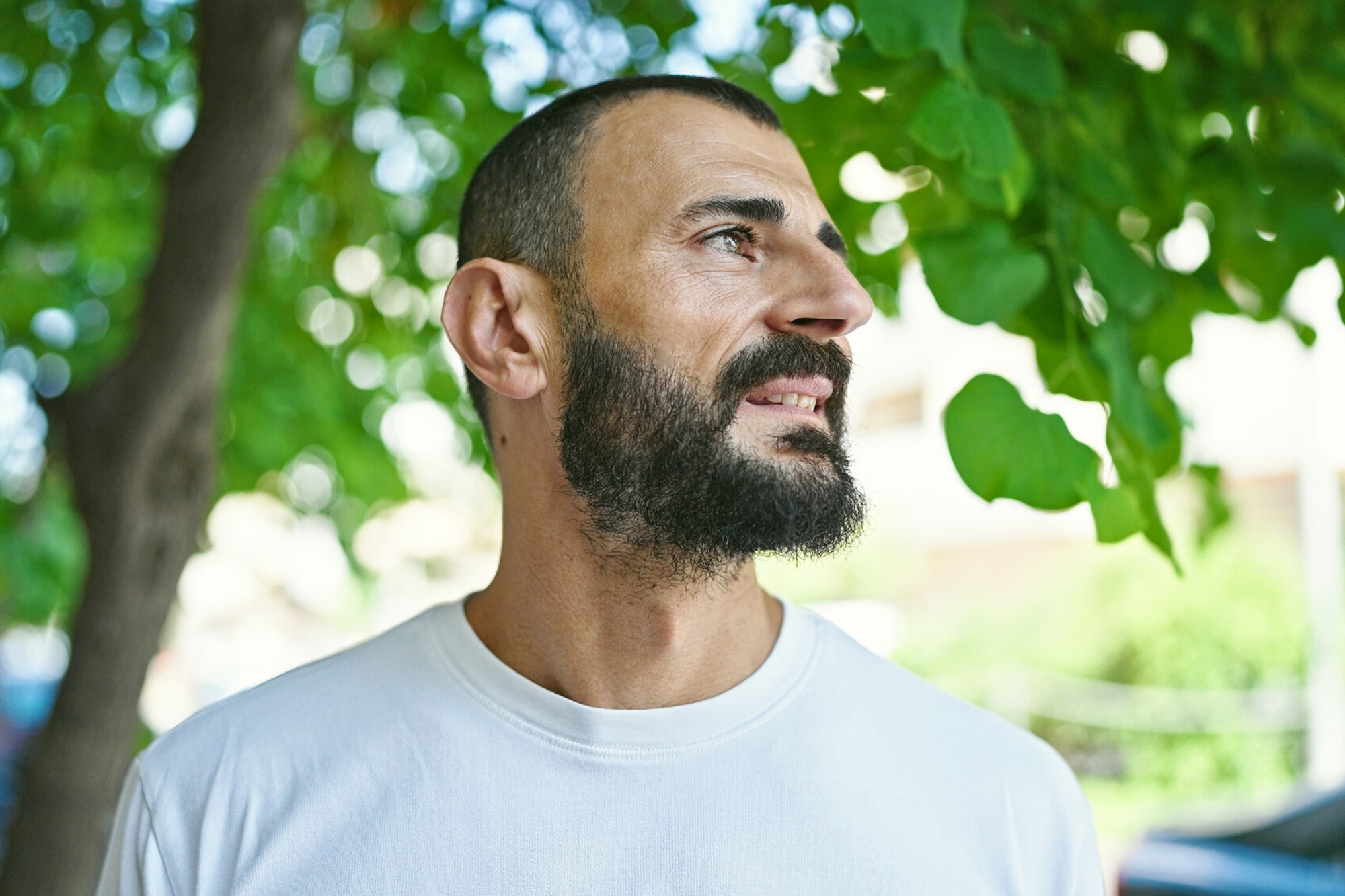 A bearded man in a white t-shirt stands outdoors, looking to his right. Green leaves and tree branches create a natural background, with sunlight filtering through.