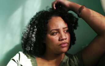 A woman with curly hair sits in soft sunlight, leaning against a green wall with her arm resting on her head, looking thoughtful and slightly concerned.