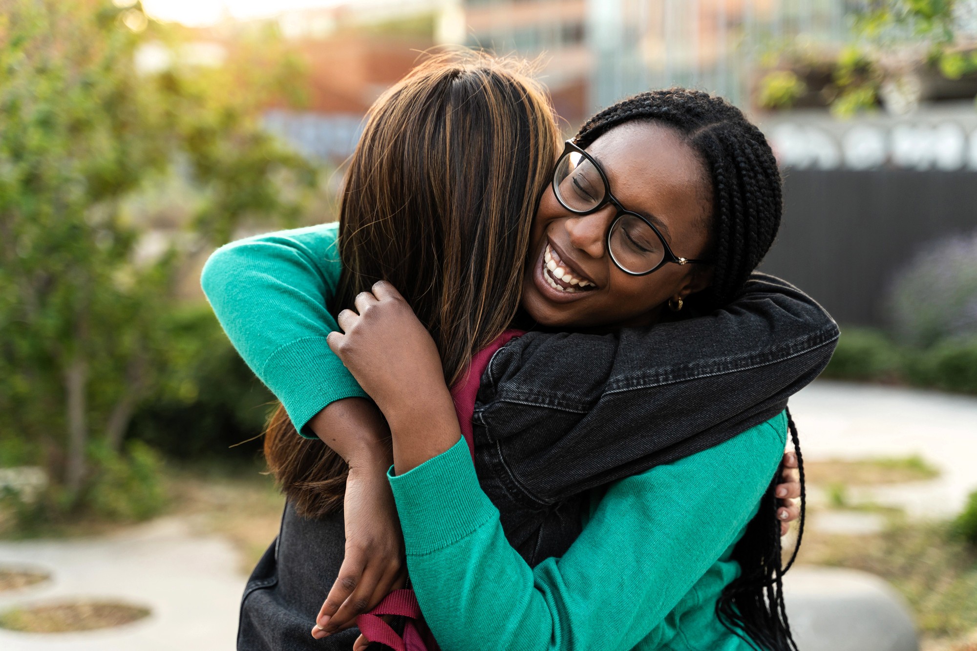Two young women embrace outdoors, smiling warmly. One has long dark braids and glasses, while the other has straight brown hair. Greenery and buildings are blurred in the background.