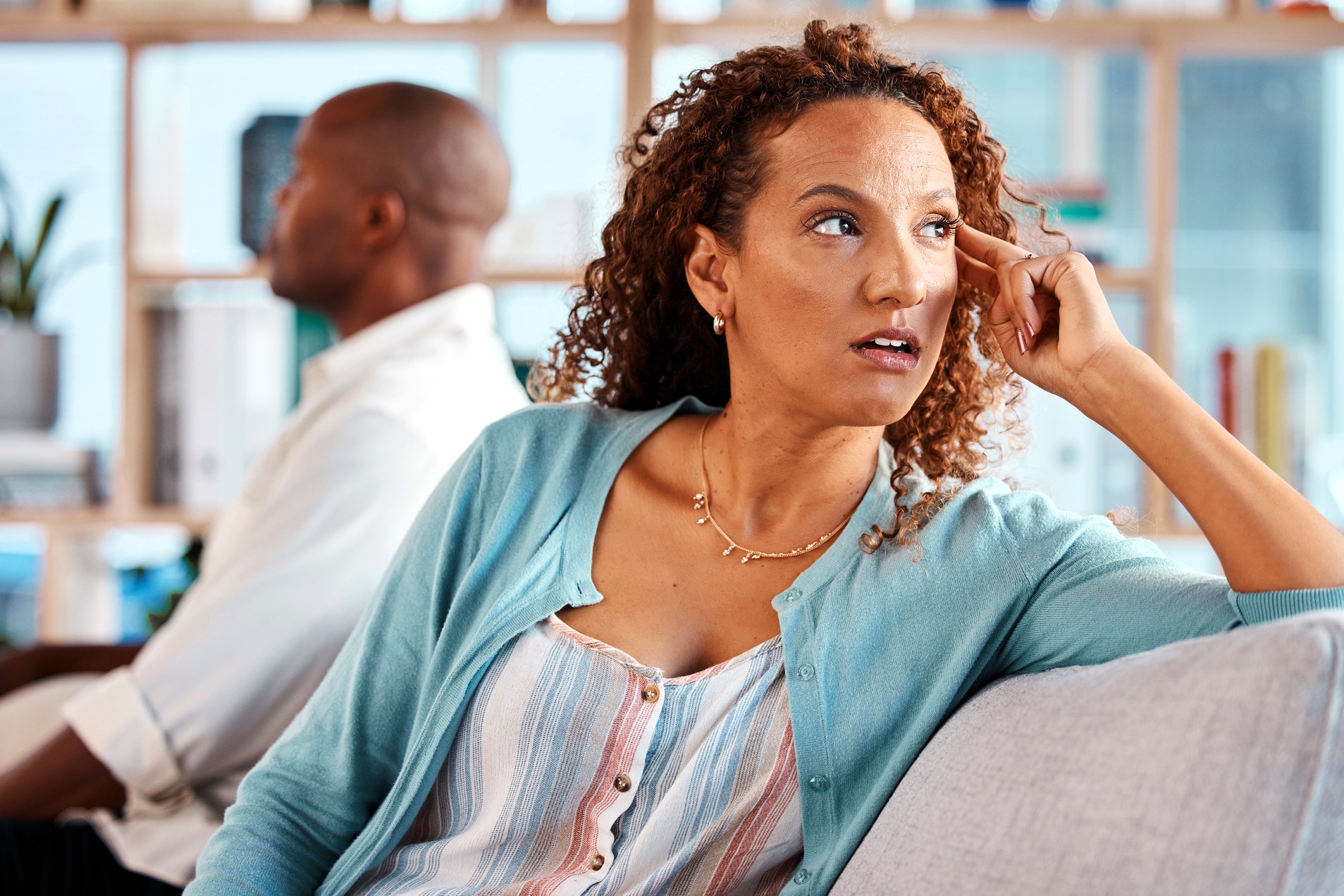 A woman with curly hair sits on a couch, looking away with a concerned expression, while a man sits in the background facing the opposite direction, both appearing upset or distant.