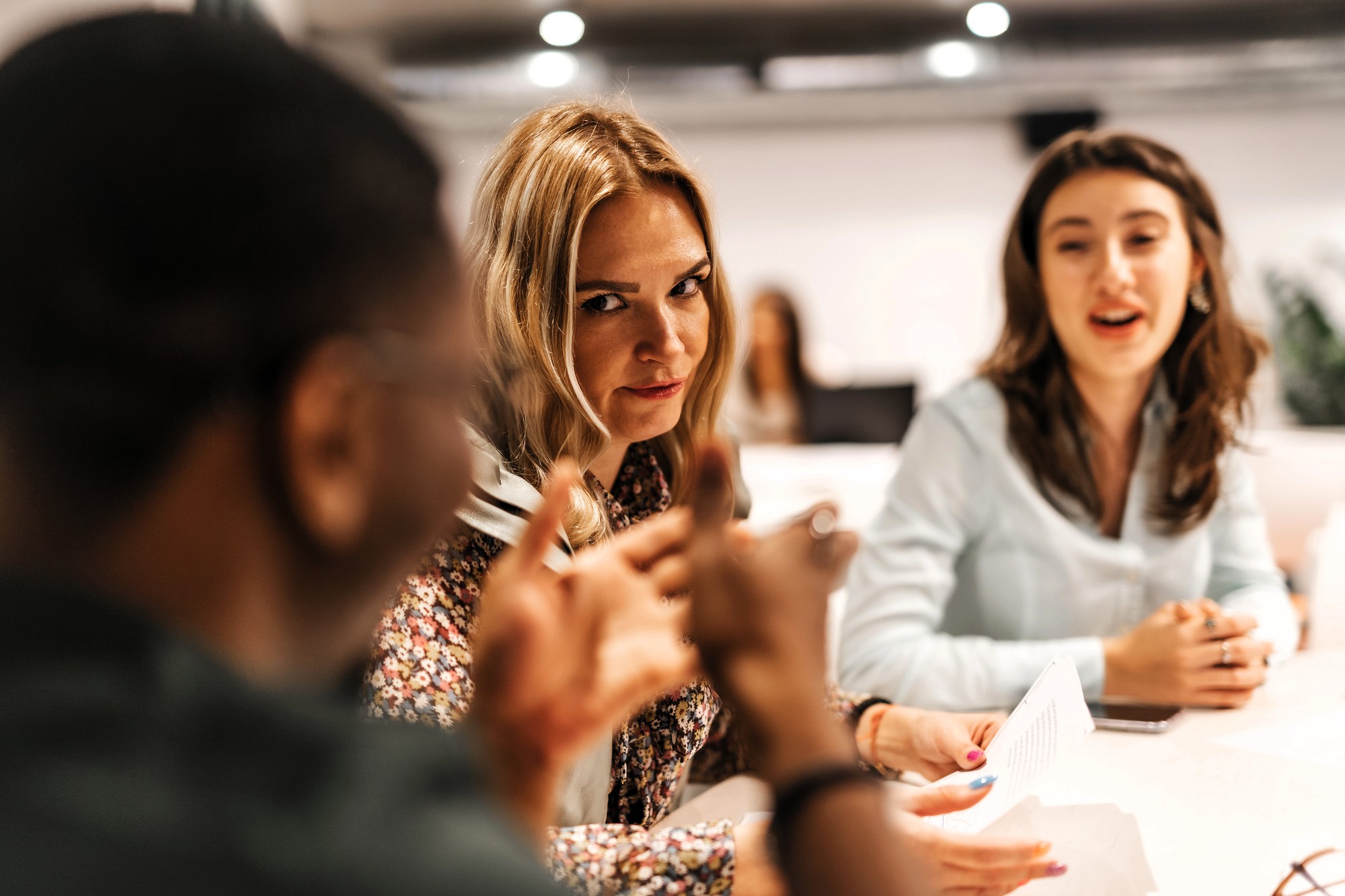 Three people sit at a table in a meeting. A woman with blonde hair in the center looks directly at the camera while listening to a man in the foreground. Another woman in the background is talking and holding papers.