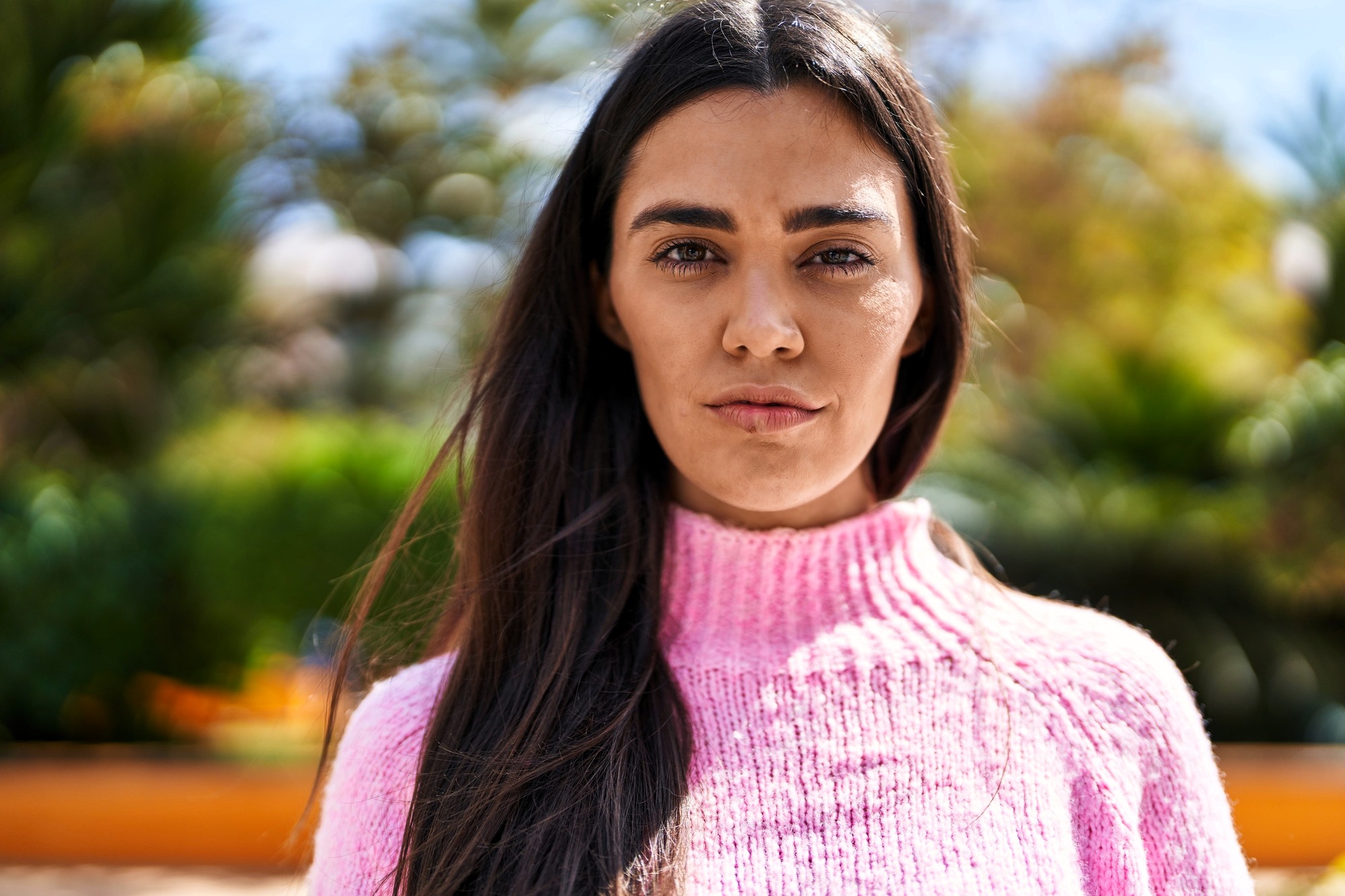 A woman with long dark hair wearing a pink sweater stands outdoors, looking directly at the camera. The background is blurred with green foliage and sunlight.