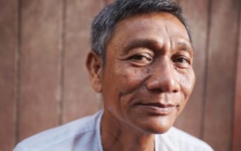 A middle-aged man with short gray hair and a slight smile looks at the camera, standing in front of a wooden background and wearing a light-colored shirt.