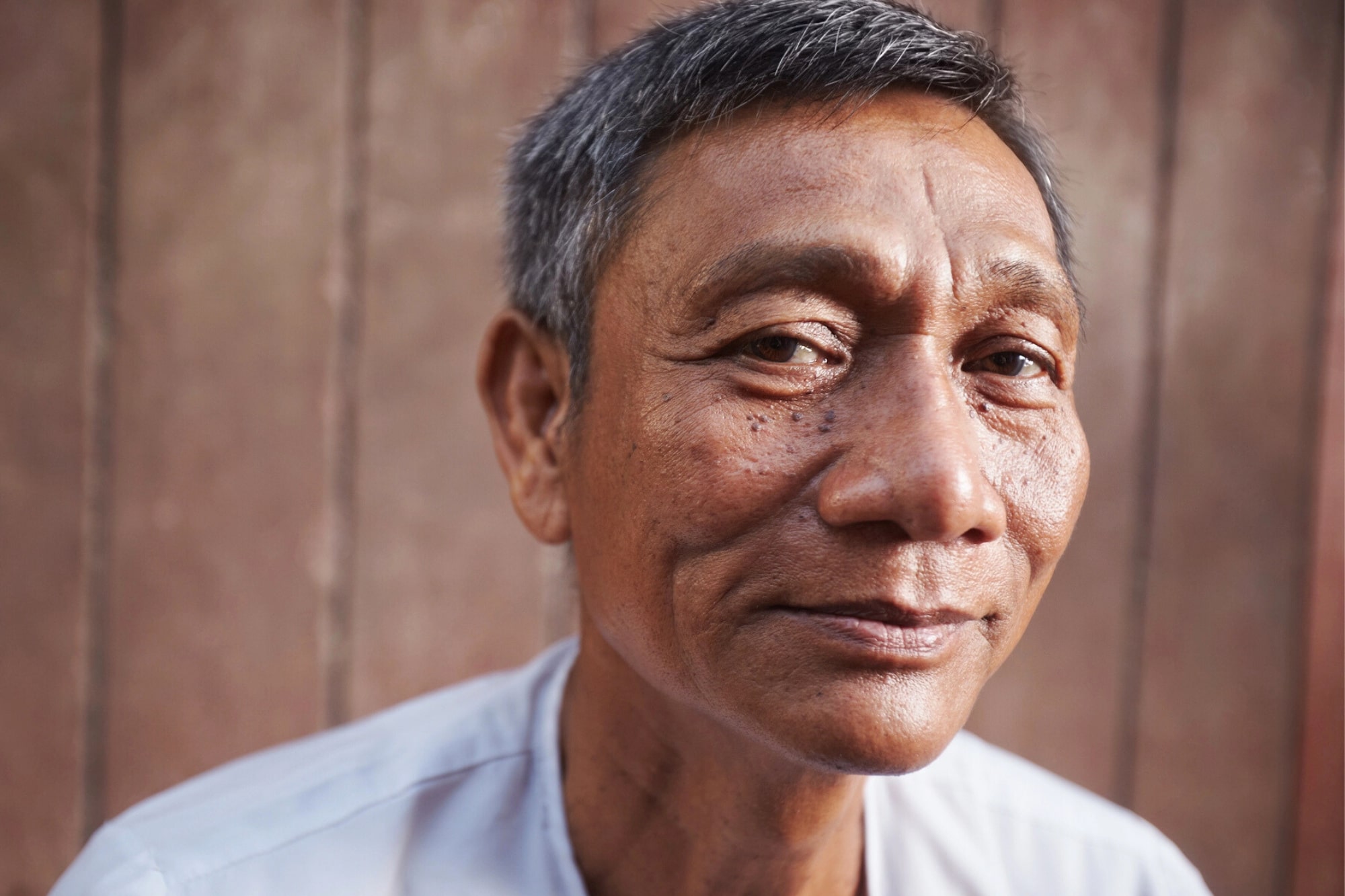 A middle-aged man with short gray hair and a slight smile looks at the camera, standing in front of a wooden background and wearing a light-colored shirt.
