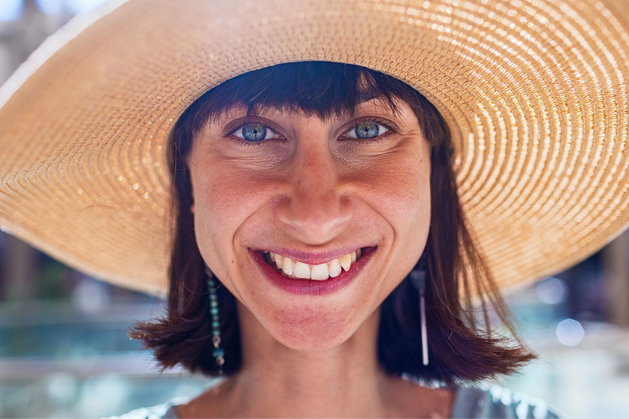 A woman with short brown hair smiles brightly, wearing a large, wide-brimmed straw hat. Sunlight highlights her face and blue eyes. The background is softly blurred, suggesting an outdoor setting.