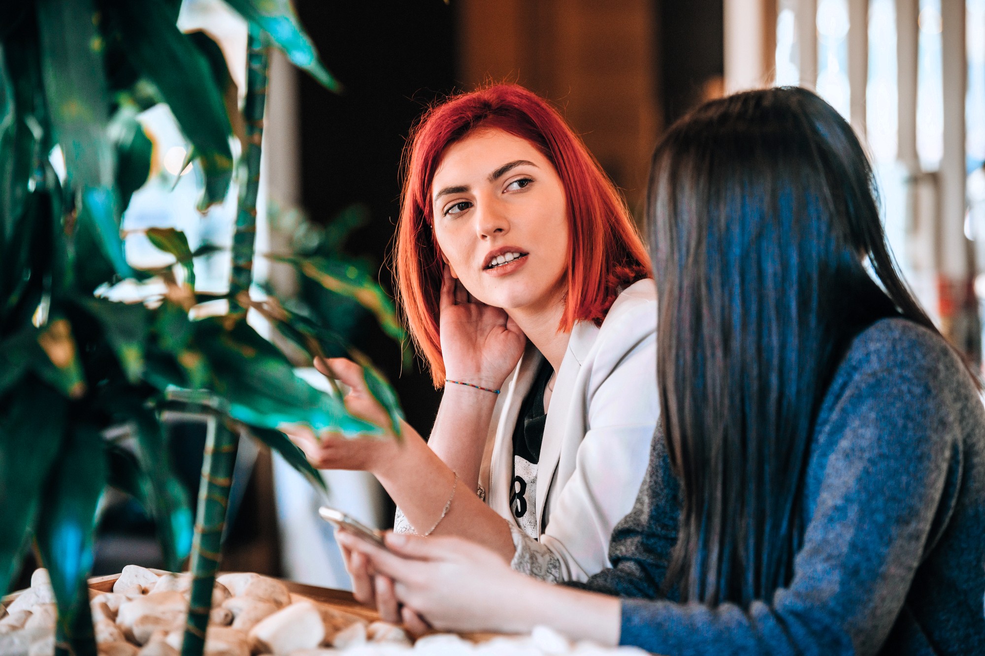 Two women are sitting indoors at a table, engaged in conversation. One has red hair and gestures while speaking; the other has long dark hair and holds a smartphone. Green plants are visible in the foreground.