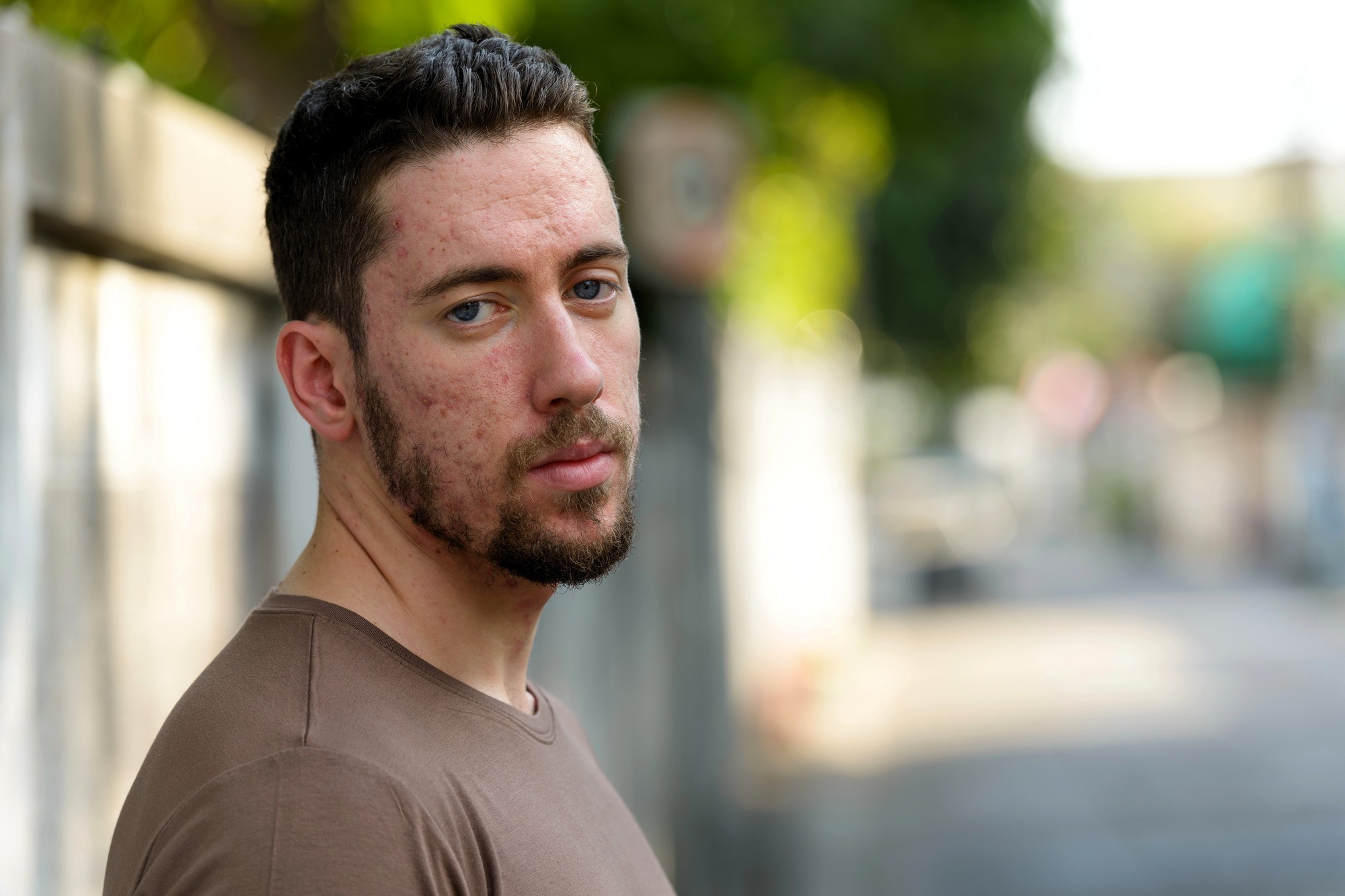 A young man with short dark hair and a trimmed beard stands outdoors, wearing a brown shirt. He looks at the camera with a neutral expression. The background is blurred, showing a street and greenery.
