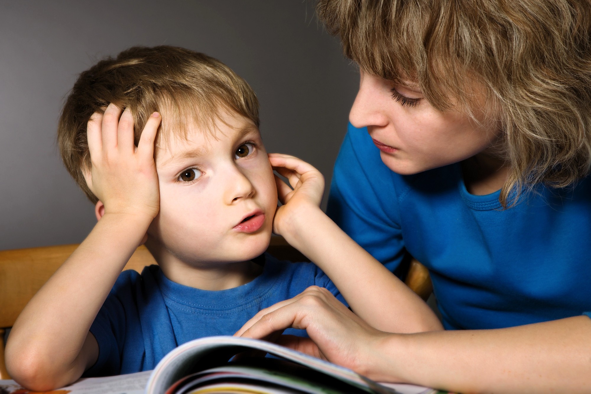 A young boy looks frustrated while sitting at a table with an open book. An adult beside him, wearing a blue shirt, offers support, gently touching his arm and looking at him with concern.