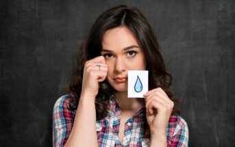 A young woman in a plaid shirt stands in front of a chalkboard, holding a paper with a blue teardrop drawn on it. She makes a sad face and gestures as if wiping a tear from her eye.