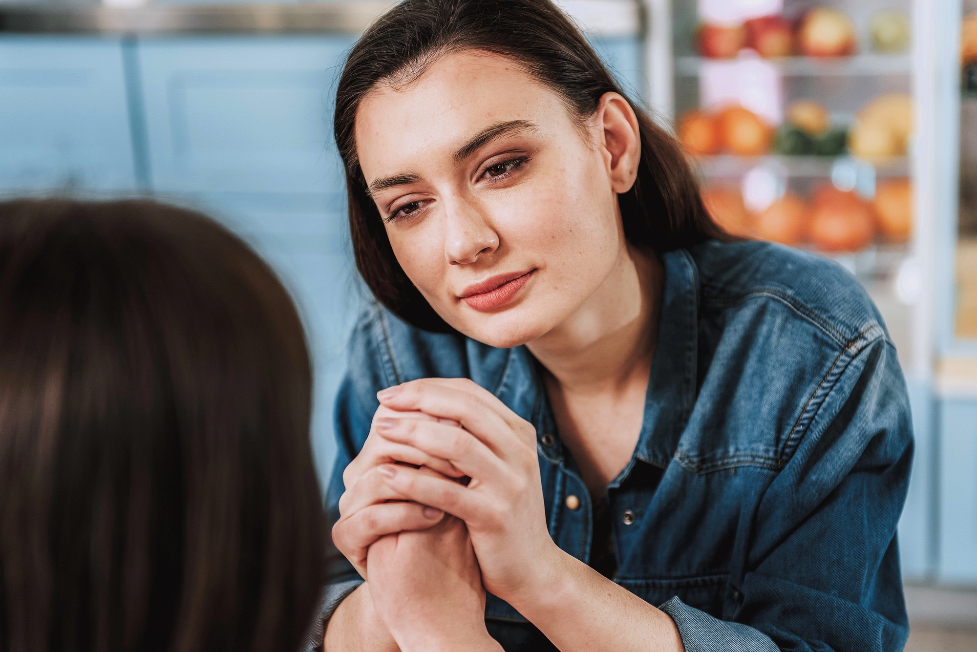 A woman in a denim shirt holds another person's hands and looks at them with a gentle, supportive expression, sitting in a kitchen with fruit visible in the background.