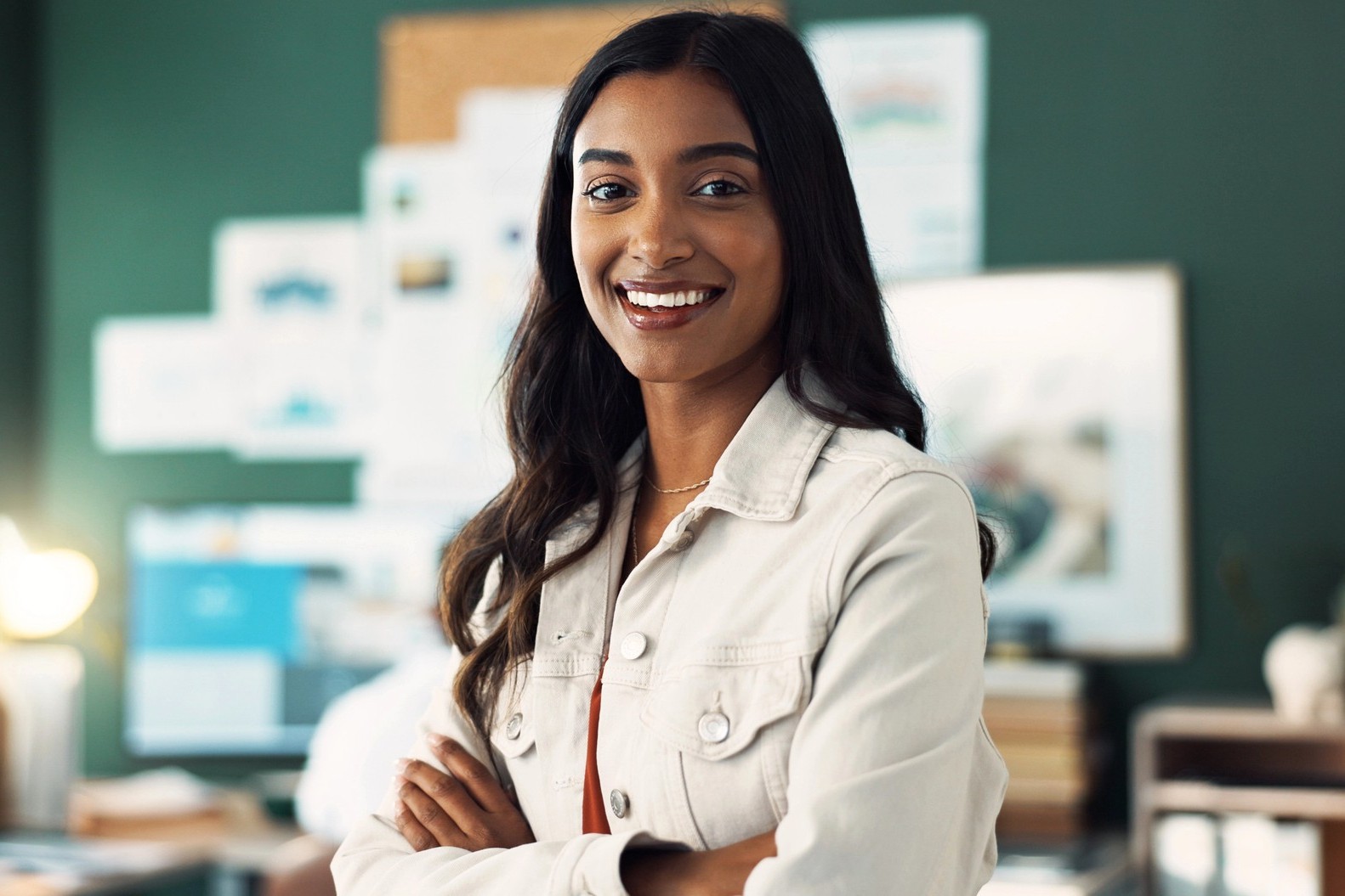 A woman with long dark hair and a light-colored jacket smiles confidently with her arms crossed, standing in an office with a dark green wall and blurred bulletin board in the background.