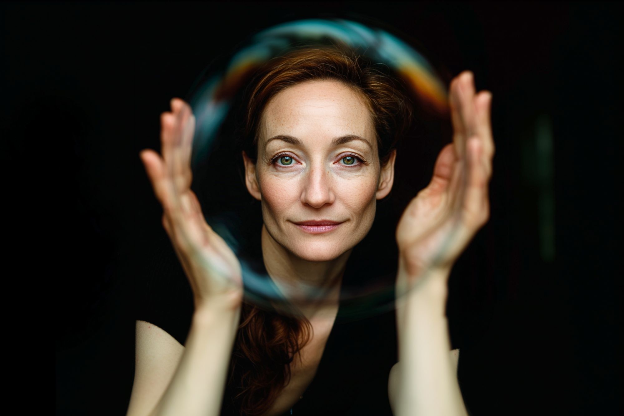 A woman with auburn hair gazes forward, holding her hands up around a large, translucent soap bubble against a dark background. The bubble appears to frame her face.
