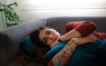A woman with long brown hair and a tattooed arm lies on a gray couch, hugging a teal pillow and looking thoughtfully ahead. Soft daylight filters in through a window behind her.
