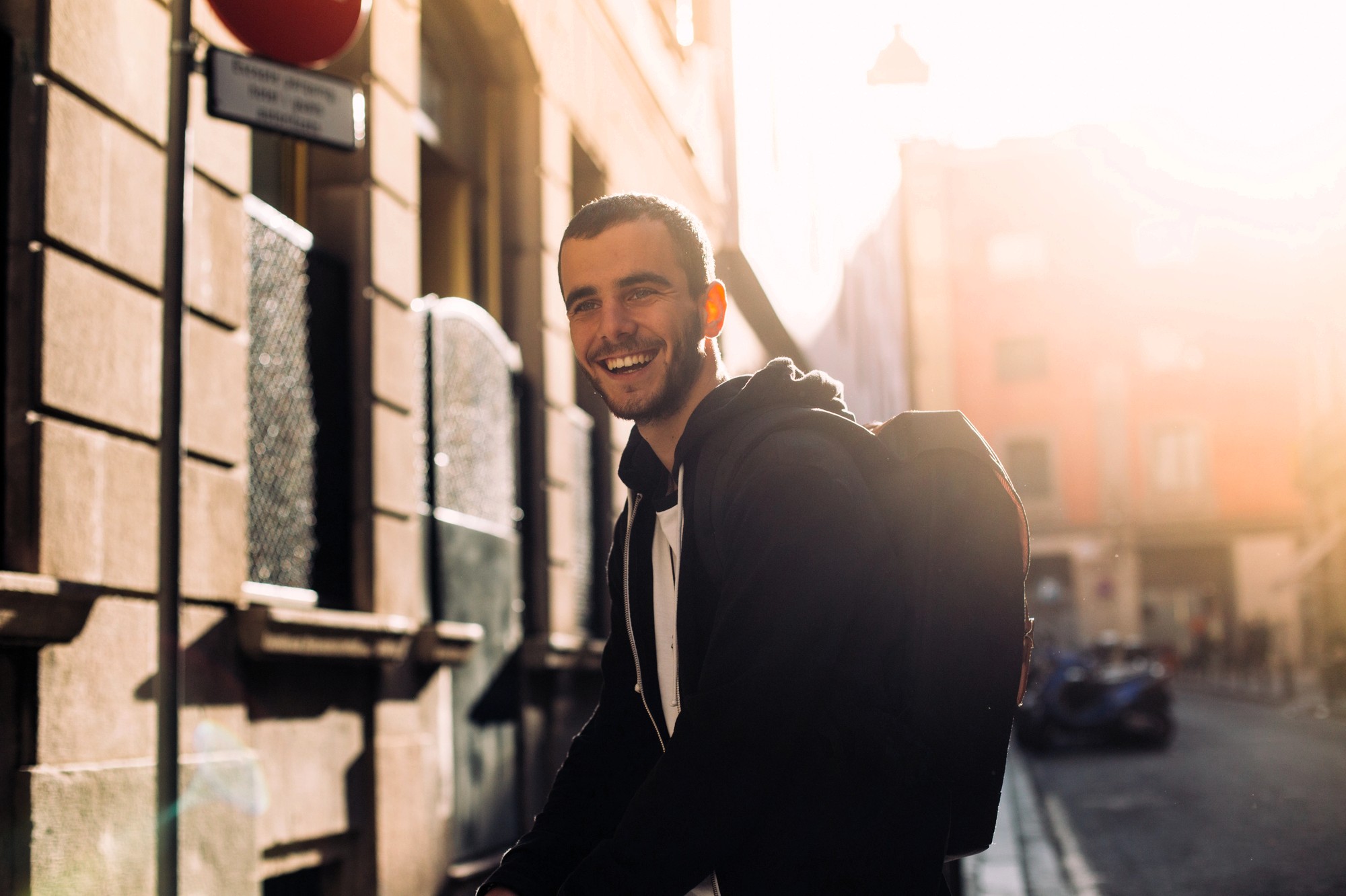 A smiling man with a backpack stands on a sunlit city street, wearing a black hoodie. Warm sunlight creates a bright glow and long shadows on the buildings and street in the background.