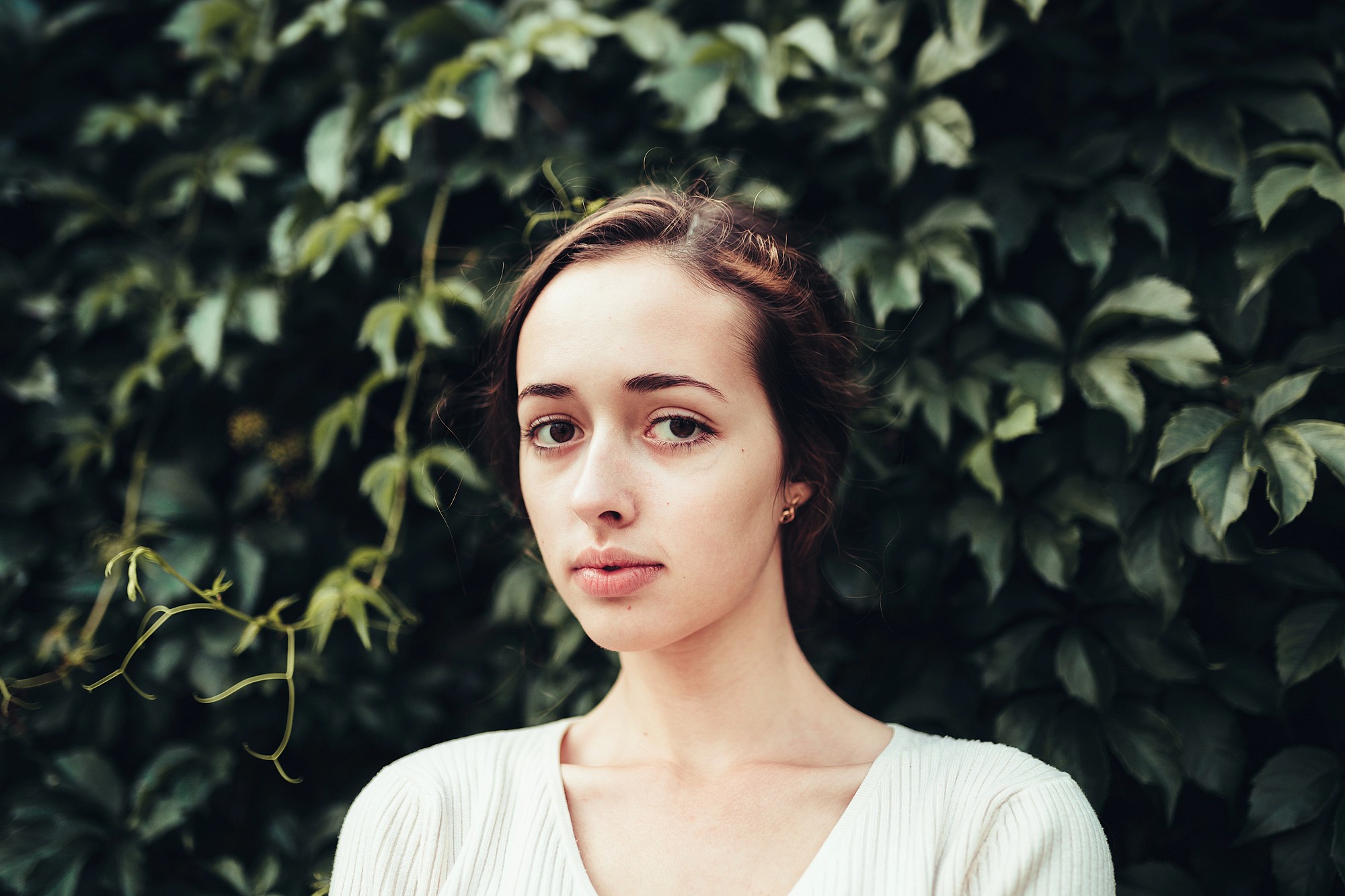 A young woman with light skin and brown hair stands in front of a wall covered in green leafy plants, looking directly at the camera with a neutral expression. She is wearing a white top.