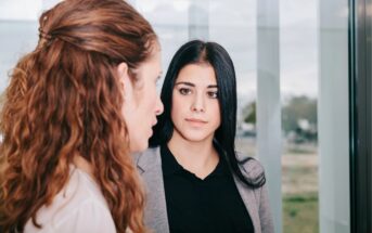 Two women are having a serious conversation indoors. One woman with long brown hair is facing away from the camera, while the other woman with straight black hair and a gray blazer looks at her attentively.