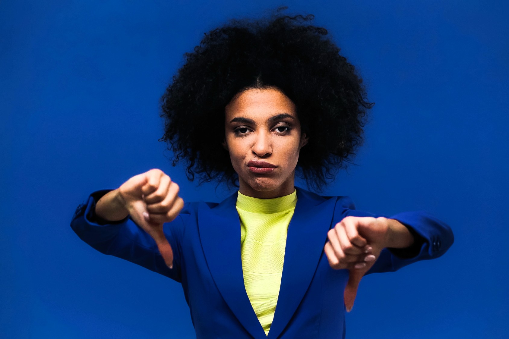 A woman with curly hair, wearing a blue blazer and yellow top, stands against a blue background, looking serious and giving two thumbs down.