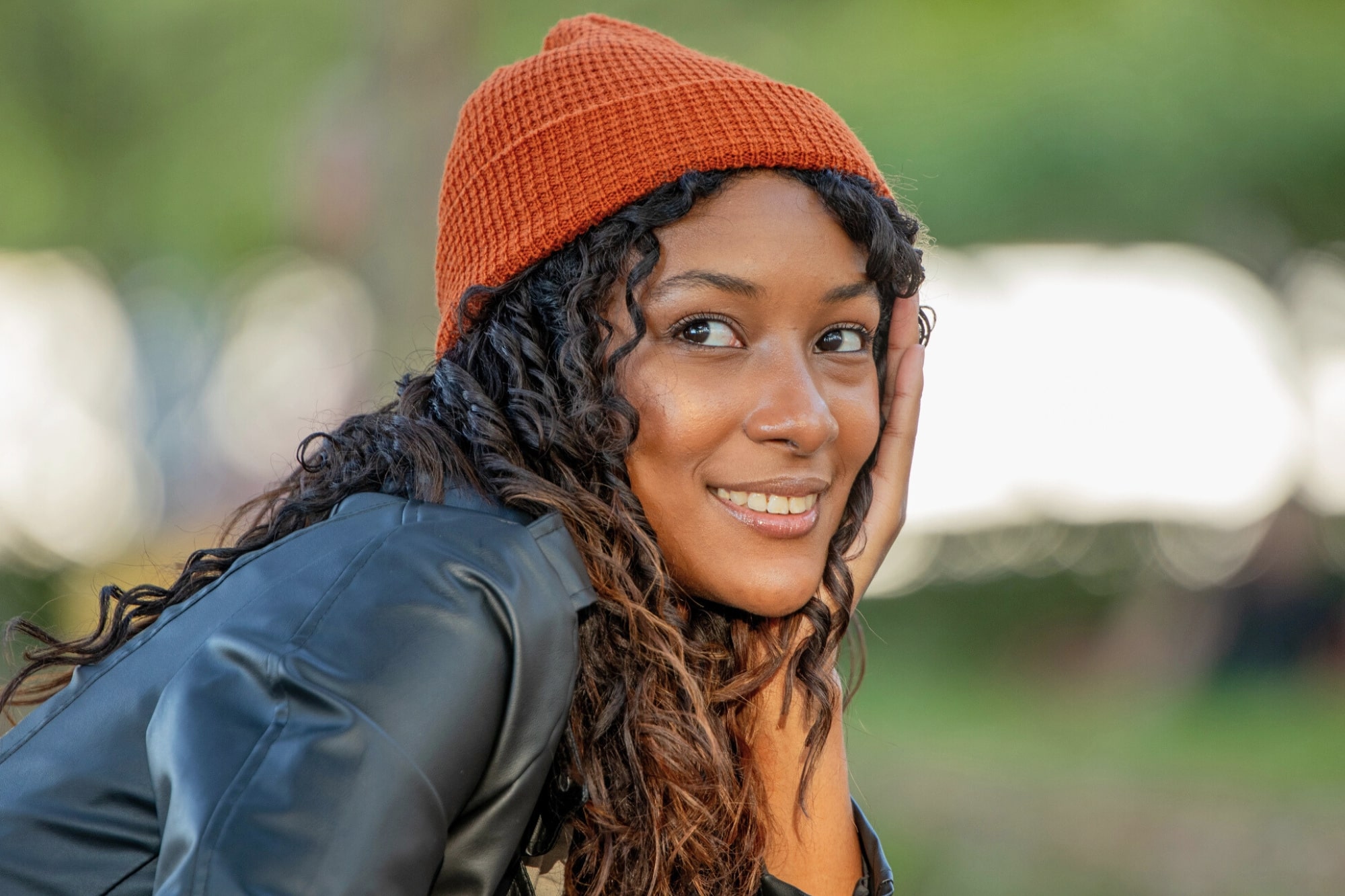 A young woman with curly hair, wearing an orange knit beanie and a black jacket, smiles while resting her face on her hand outdoors with a blurred green background.