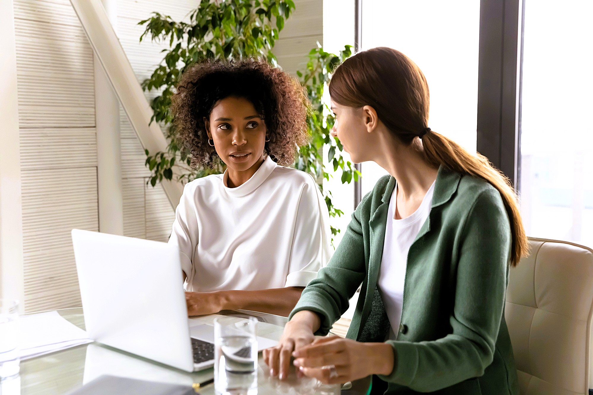 Two women sit at a desk in an office, having a conversation. One is wearing a white top and the other a green blazer. A laptop, papers, and a glass of water are on the desk, with plants and windows in the background.