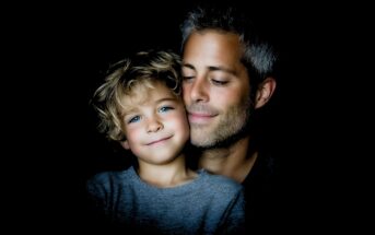A man with gray hair smiles gently, holding a young child with curly blonde hair and blue eyes. Both are facing forward against a dark background, illuminated by soft light.