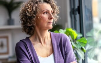 A woman with curly hair and a purple cardigan looks thoughtfully out of a window, standing indoors with her arms crossed. Green plants and soft light are visible in the background.