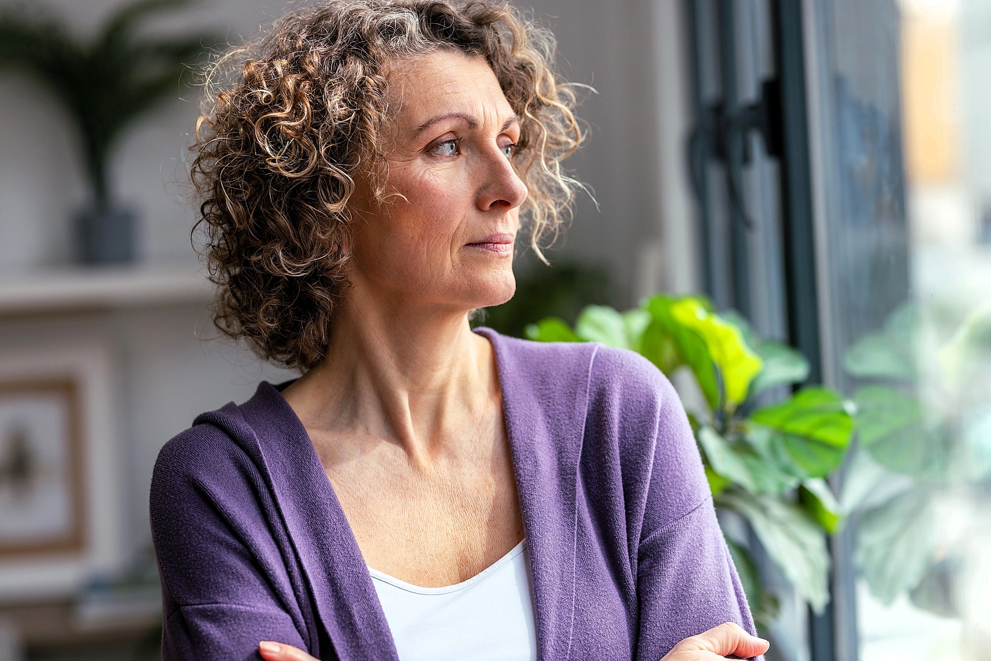 A woman with curly hair and a purple cardigan looks thoughtfully out of a window, standing indoors with her arms crossed. Green plants and soft light are visible in the background.