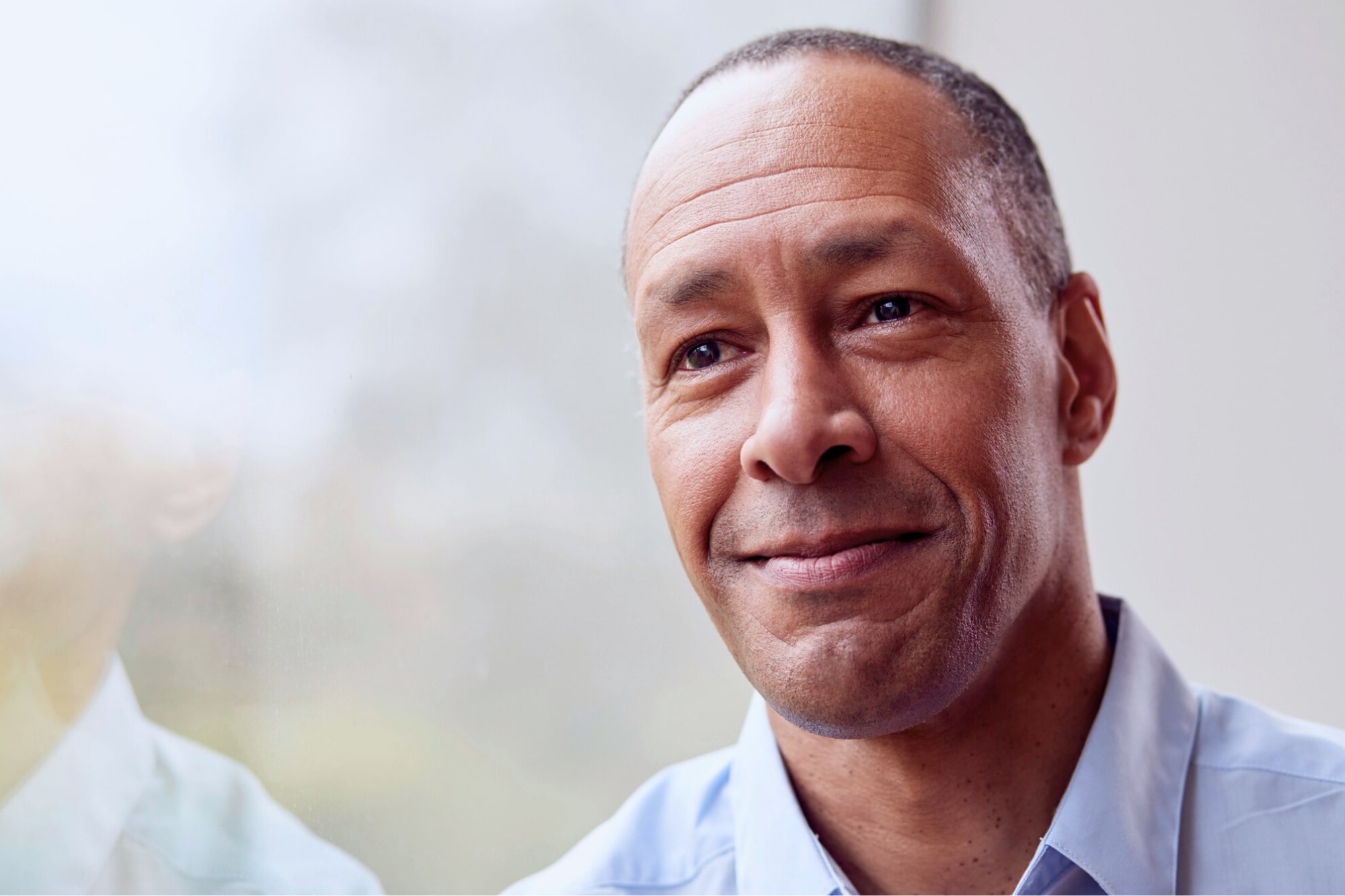 A middle-aged man with short hair and a slight smile looks thoughtfully out of a window. He is wearing a light blue shirt, and soft natural light illuminates his face.