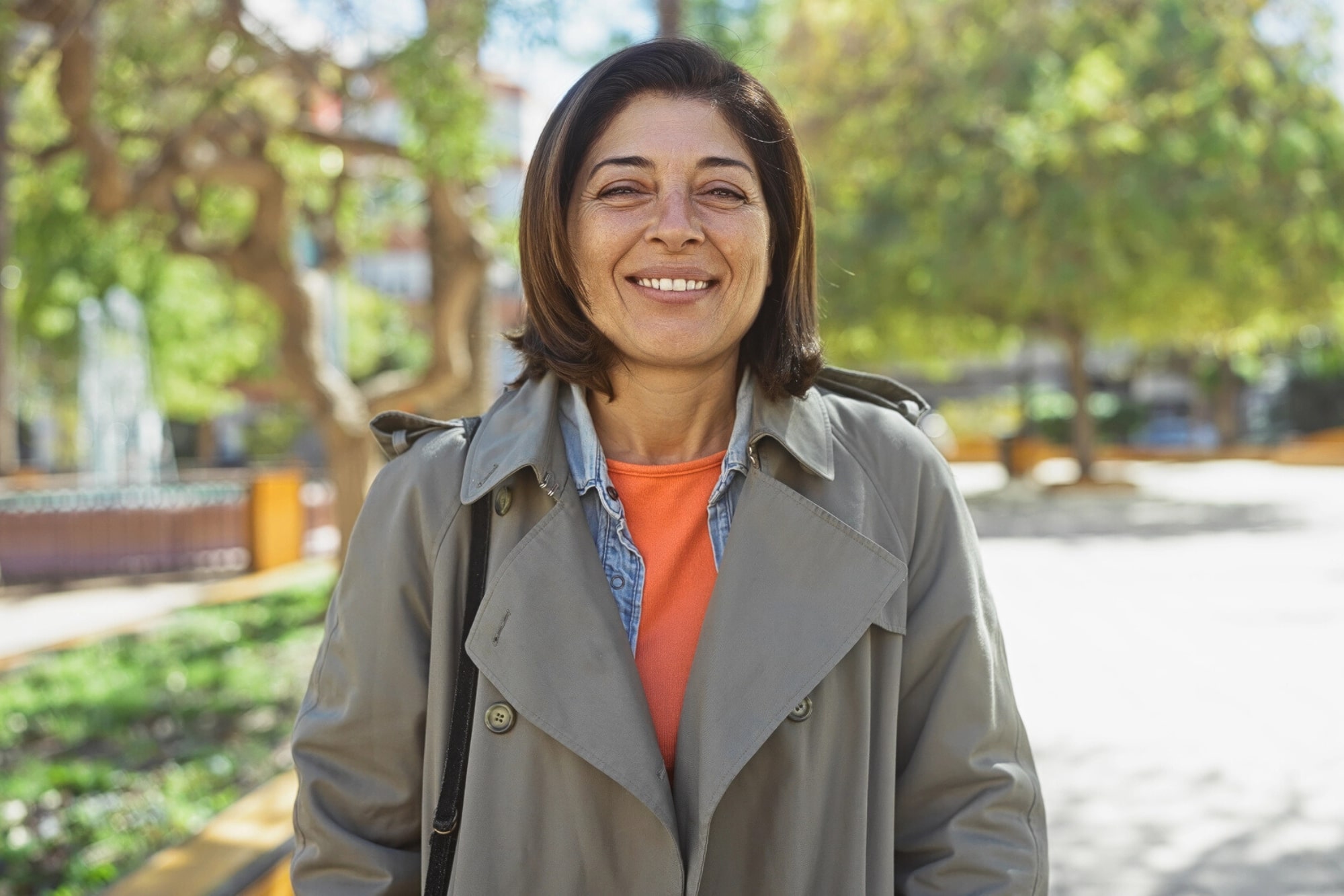 A woman with shoulder-length brown hair wearing a tan trench coat and orange top stands outdoors, smiling, with trees and sunlight in the background.