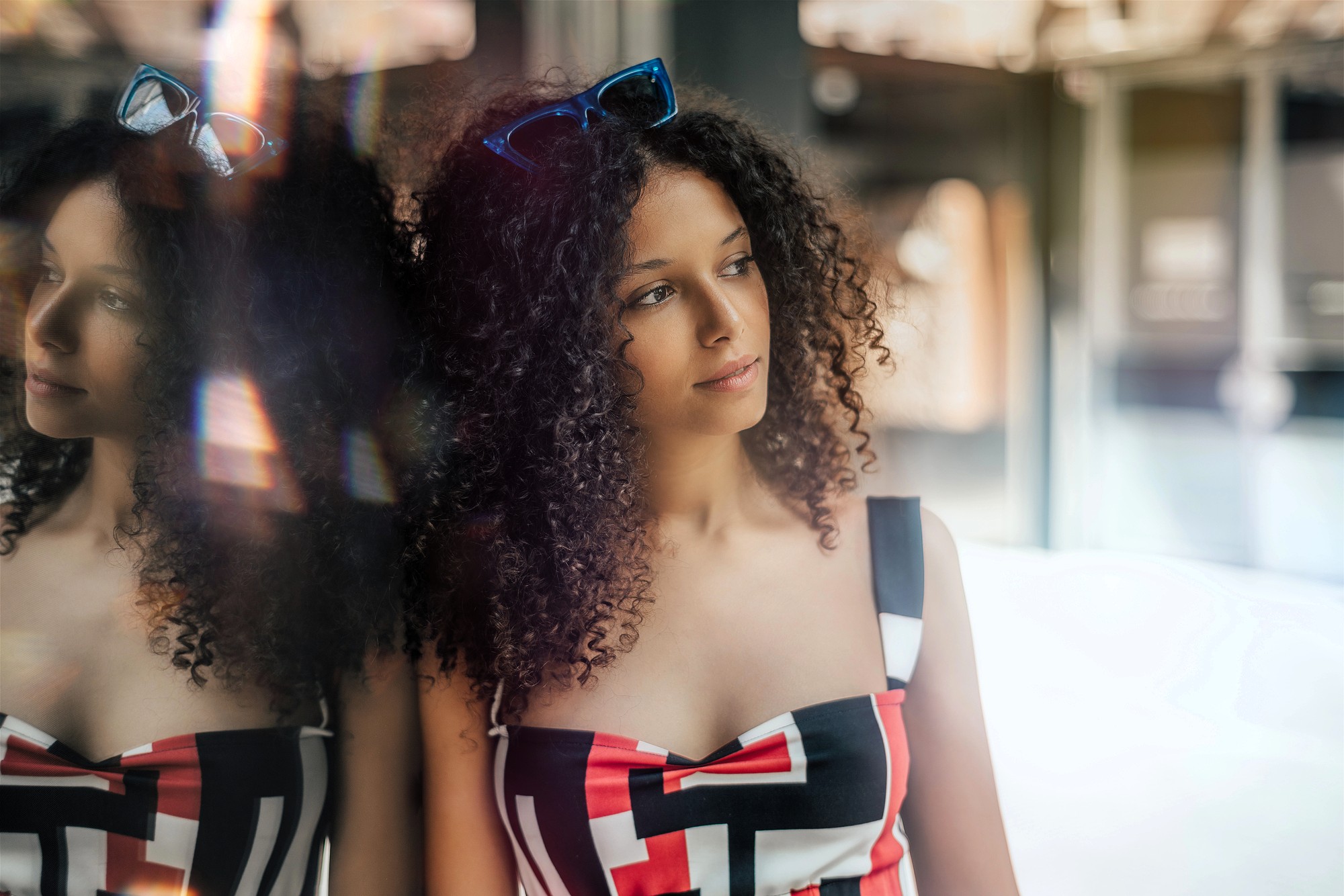 A woman with curly hair wearing a geometric-patterned top and sunglasses on her head looks thoughtfully out of frame, leaning against a window that reflects her image.