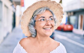 Smiling older woman with gray hair wearing glasses, a straw hat, and a white off-shoulder top stands outdoors on a sunny street, looking slightly upward.