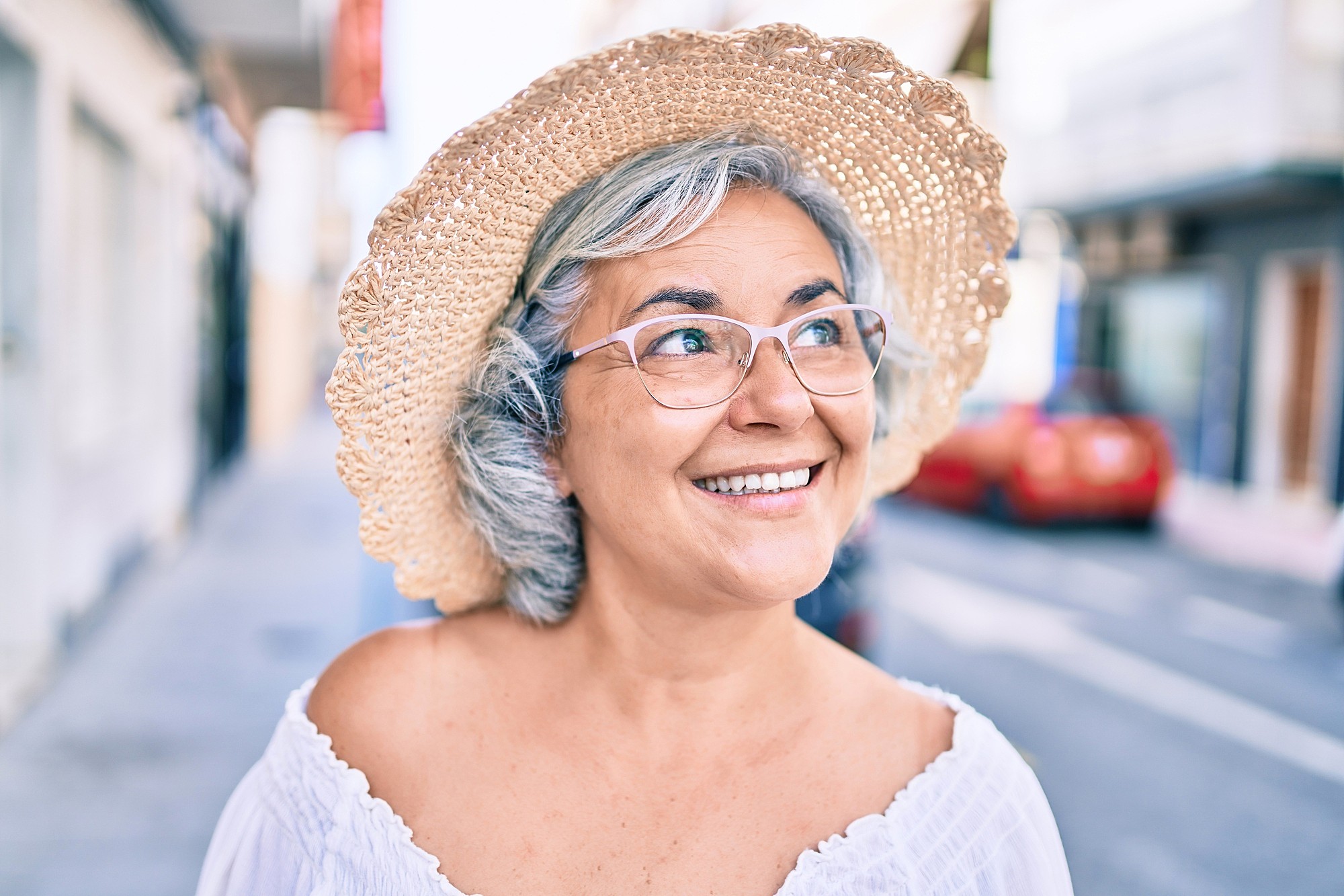 Smiling older woman with gray hair wearing glasses, a straw hat, and a white off-shoulder top stands outdoors on a sunny street, looking slightly upward.