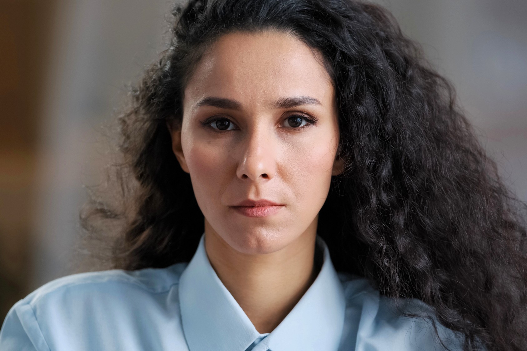 A woman with long, curly dark hair wearing a light blue collared shirt looks directly at the camera with a neutral expression. The background is softly blurred.
