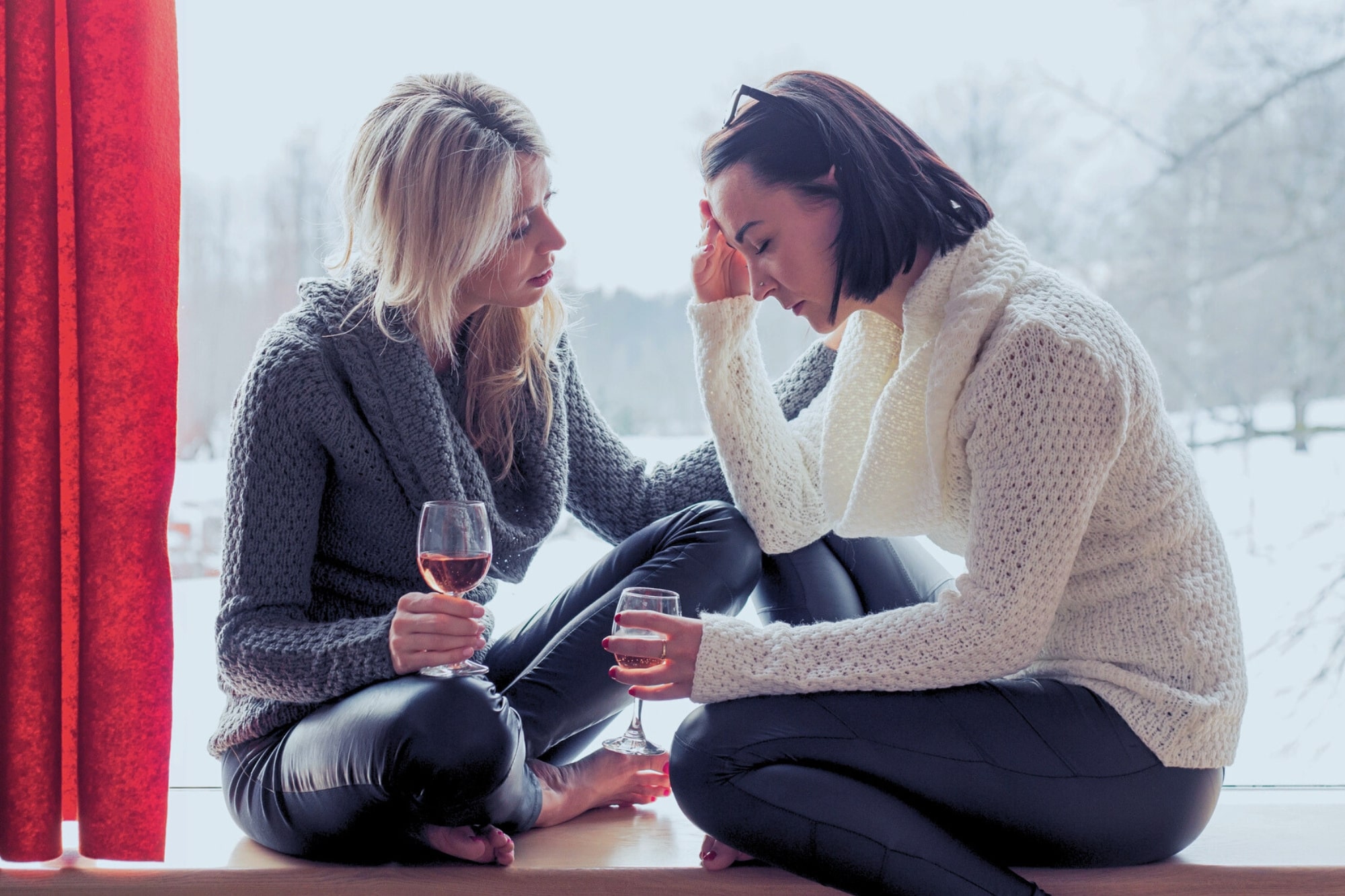 Two women in cozy sweaters sit by a window holding wine glasses. One woman appears upset with her head in her hand, while the other gently comforts her. Snow is visible outside the window.