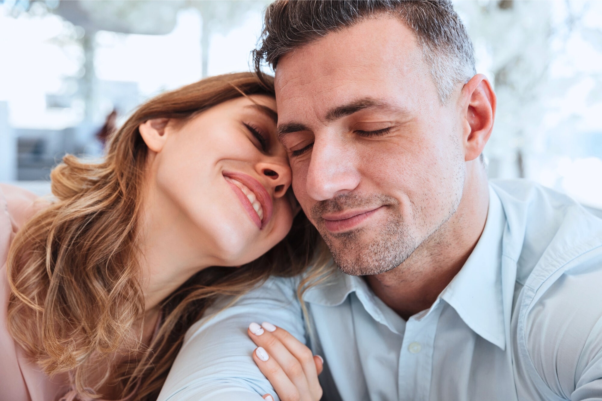 A smiling woman leans her head affectionately on a man’s shoulder. Both have their eyes closed, appearing content and relaxed. The background is softly blurred, suggesting a bright indoor setting.