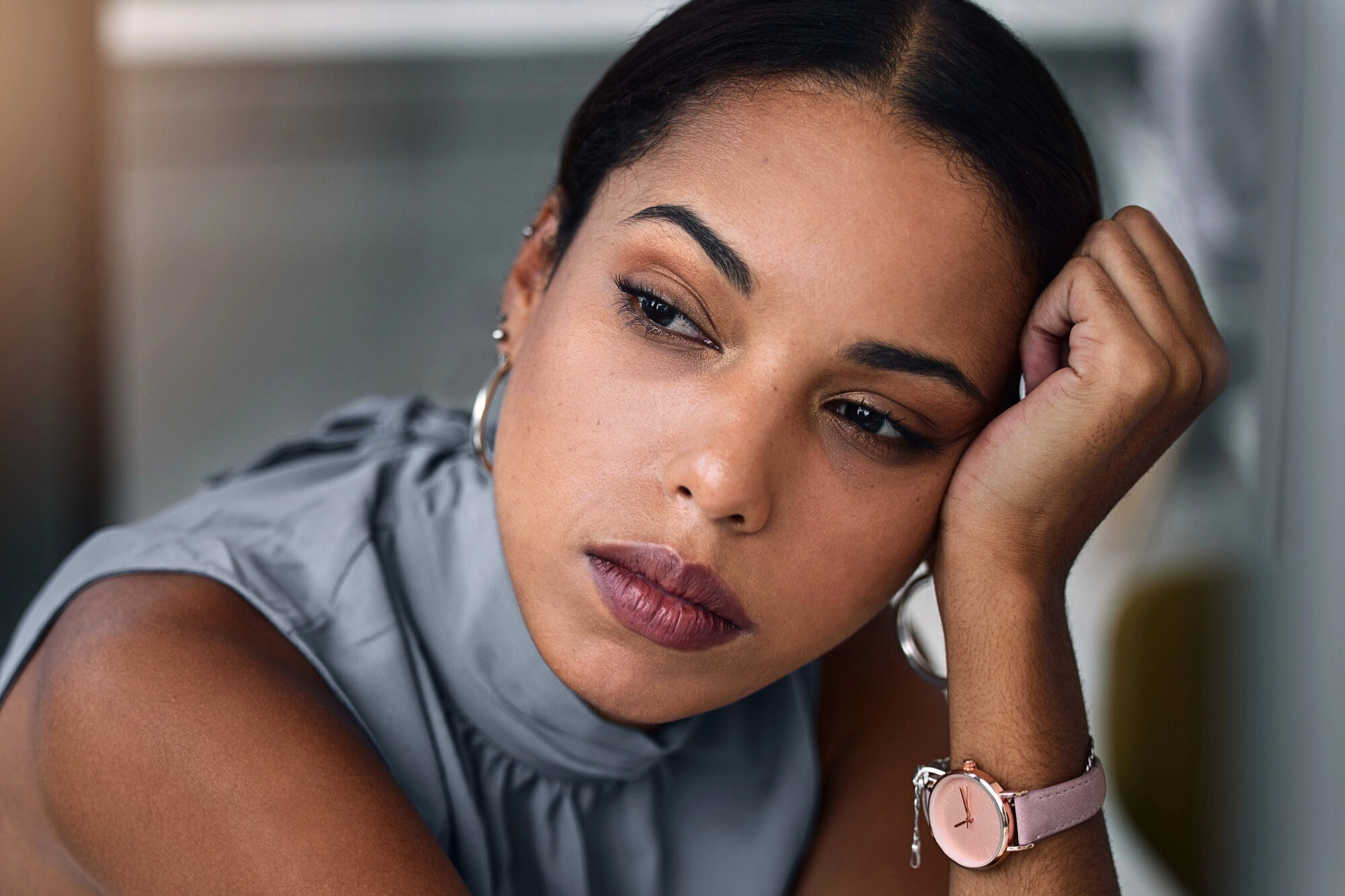 A woman with medium skin tone rests her face on her hand, looking thoughtfully into the distance. She wears a sleeveless gray top, silver hoop earrings, and a pink wristwatch.