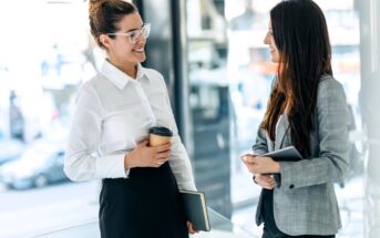 Two women in business attire stand indoors, smiling and talking. One holds a coffee cup and notebook, while the other holds a tablet. Bright, modern office environment with large windows in the background.