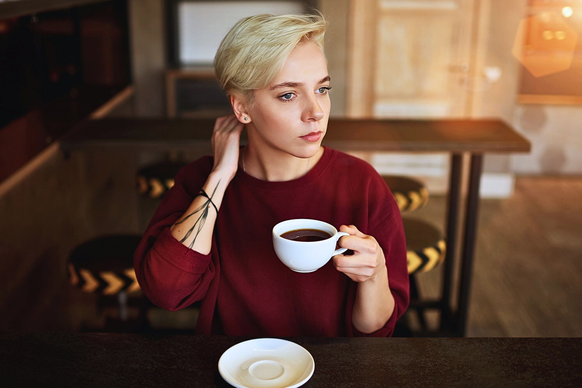 A person with short blond hair wearing a red sweater sits at a table in a café, holding a cup of coffee and looking thoughtfully into the distance. An empty saucer is on the table in front of them.