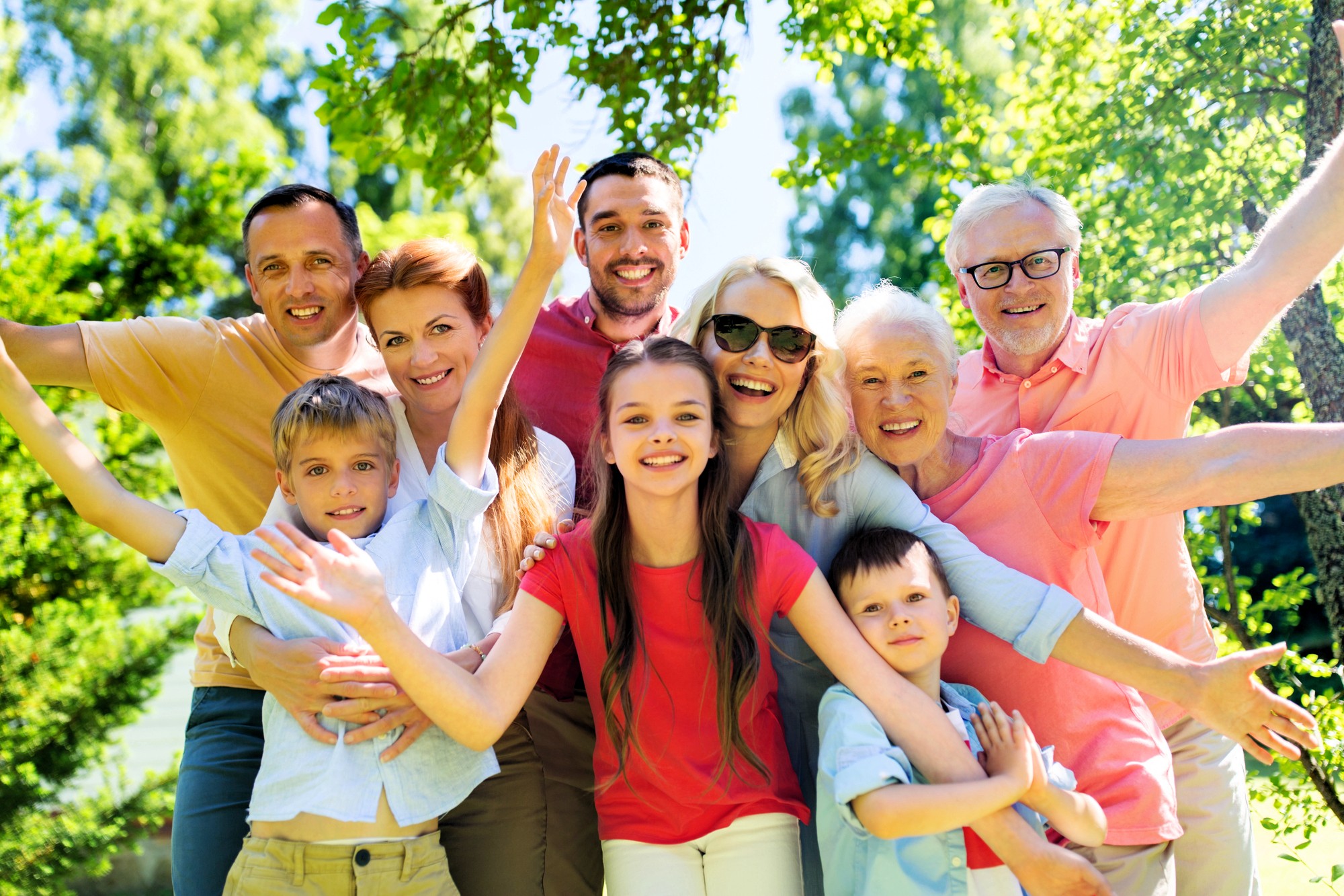 A large, happy family of multiple generations stands close together outdoors, smiling and posing with arms outstretched on a sunny day surrounded by green trees.