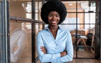 A woman with a natural afro hairstyle, wearing a light blue button-up shirt, stands confidently with arms crossed and smiles in a modern office with glass walls and wooden floors.