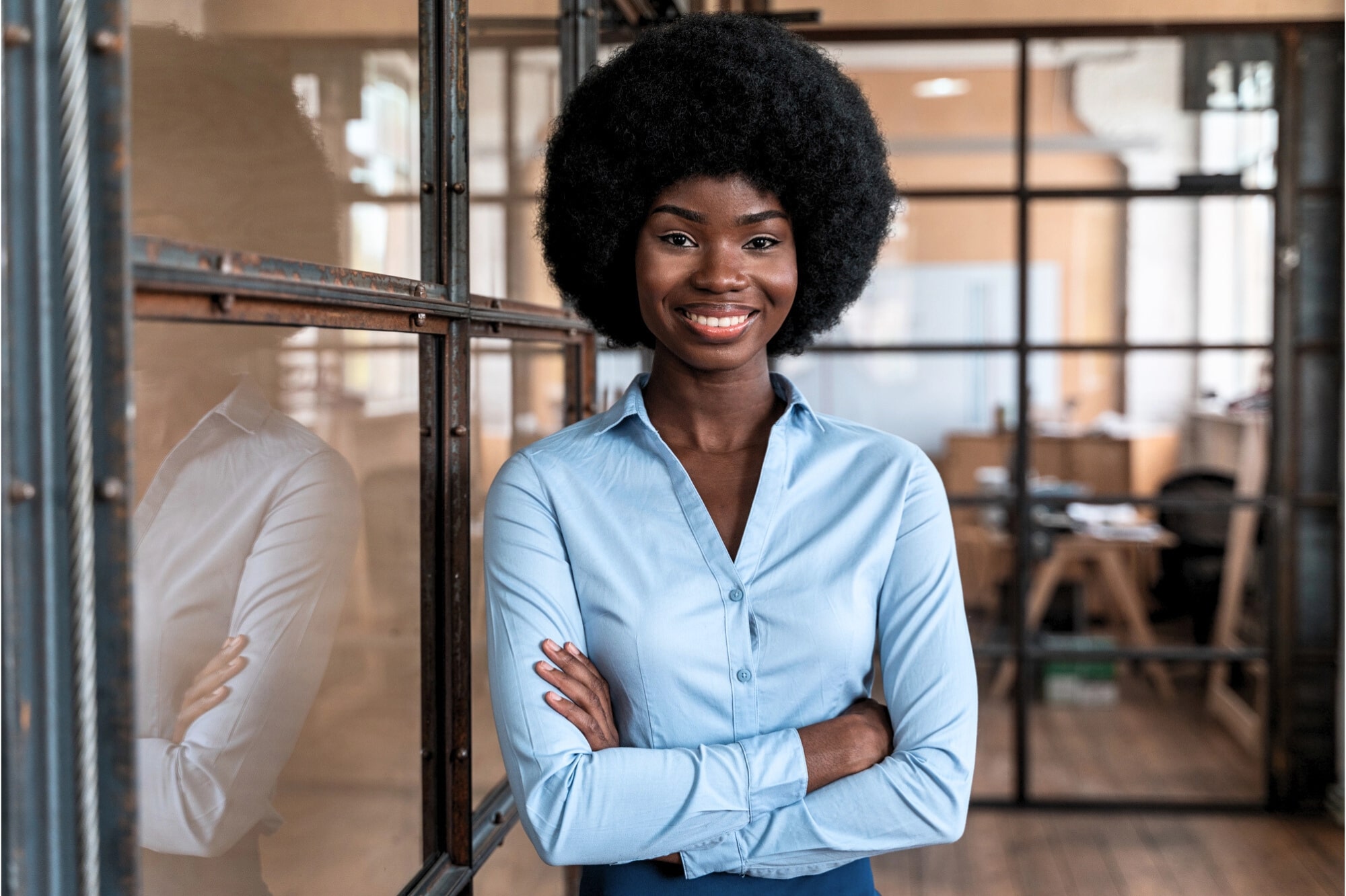 A woman with a natural afro hairstyle, wearing a light blue button-up shirt, stands confidently with arms crossed and smiles in a modern office with glass walls and wooden floors.