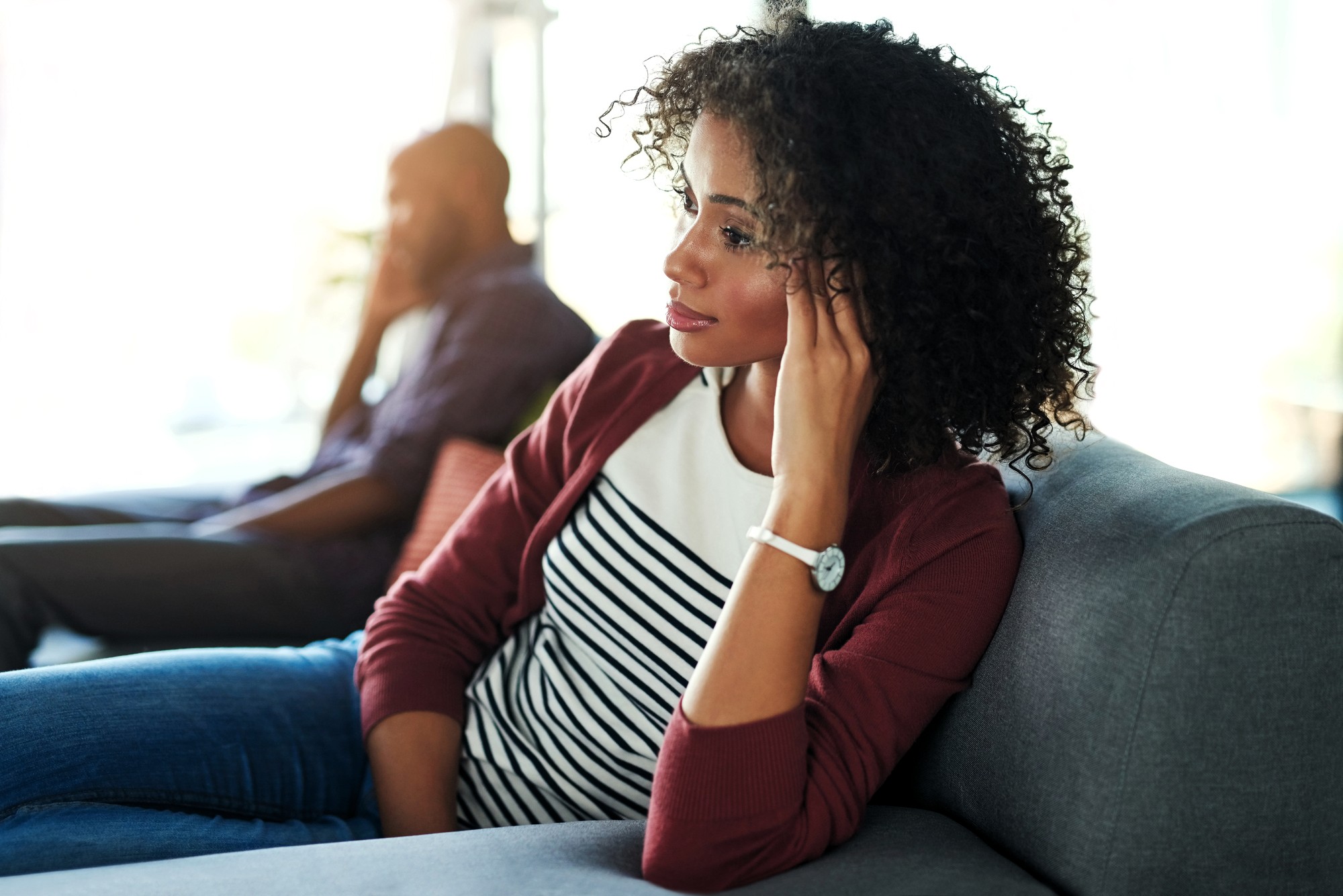 A woman with curly hair sits thoughtfully on a couch, looking away, with her hand touching her ear. A man in the background sits on another part of the couch, slightly out of focus.