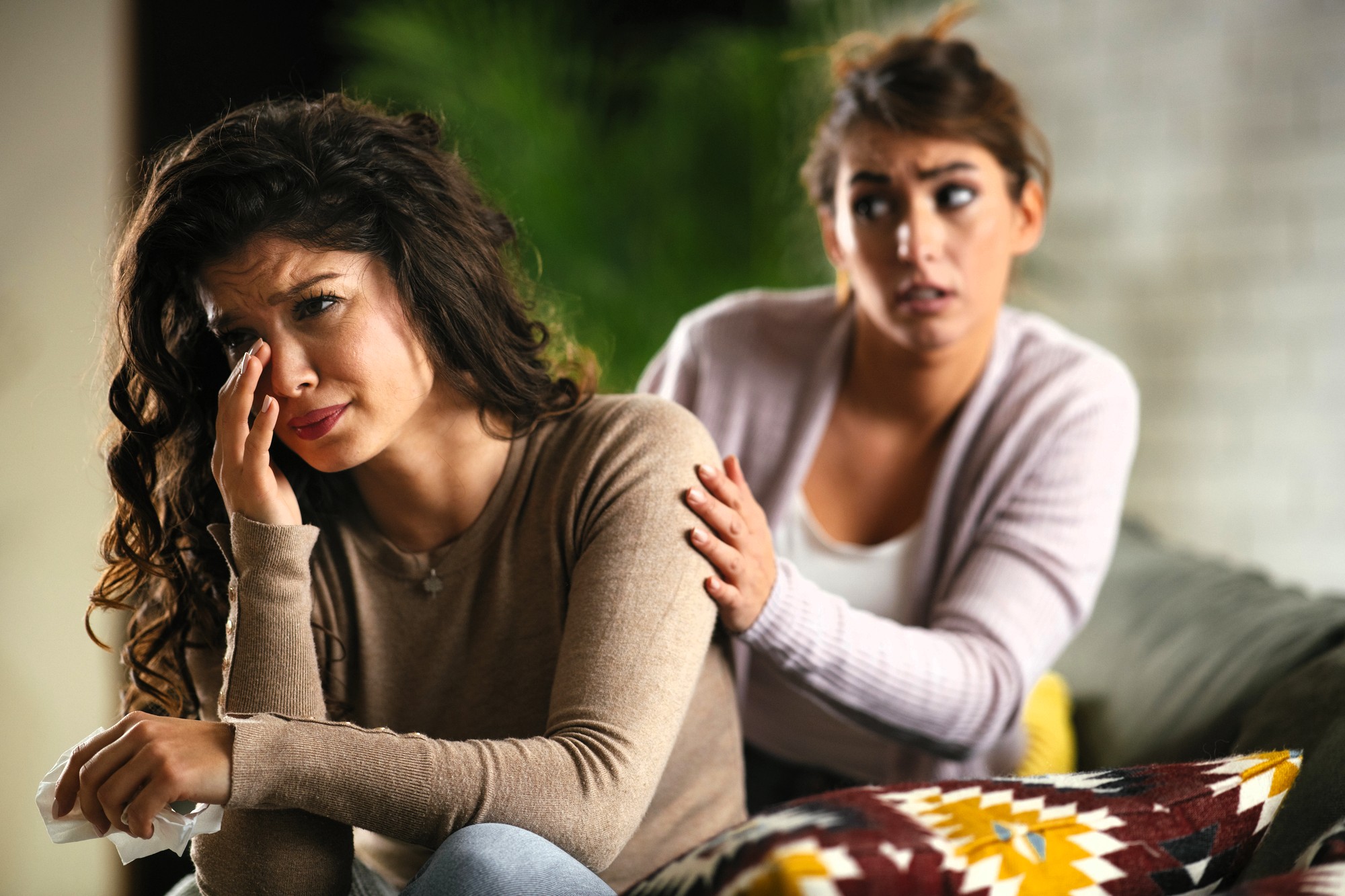 A woman sits on a couch, crying and wiping her face with a tissue, while another woman sits beside her, gently comforting her with a hand on her shoulder.