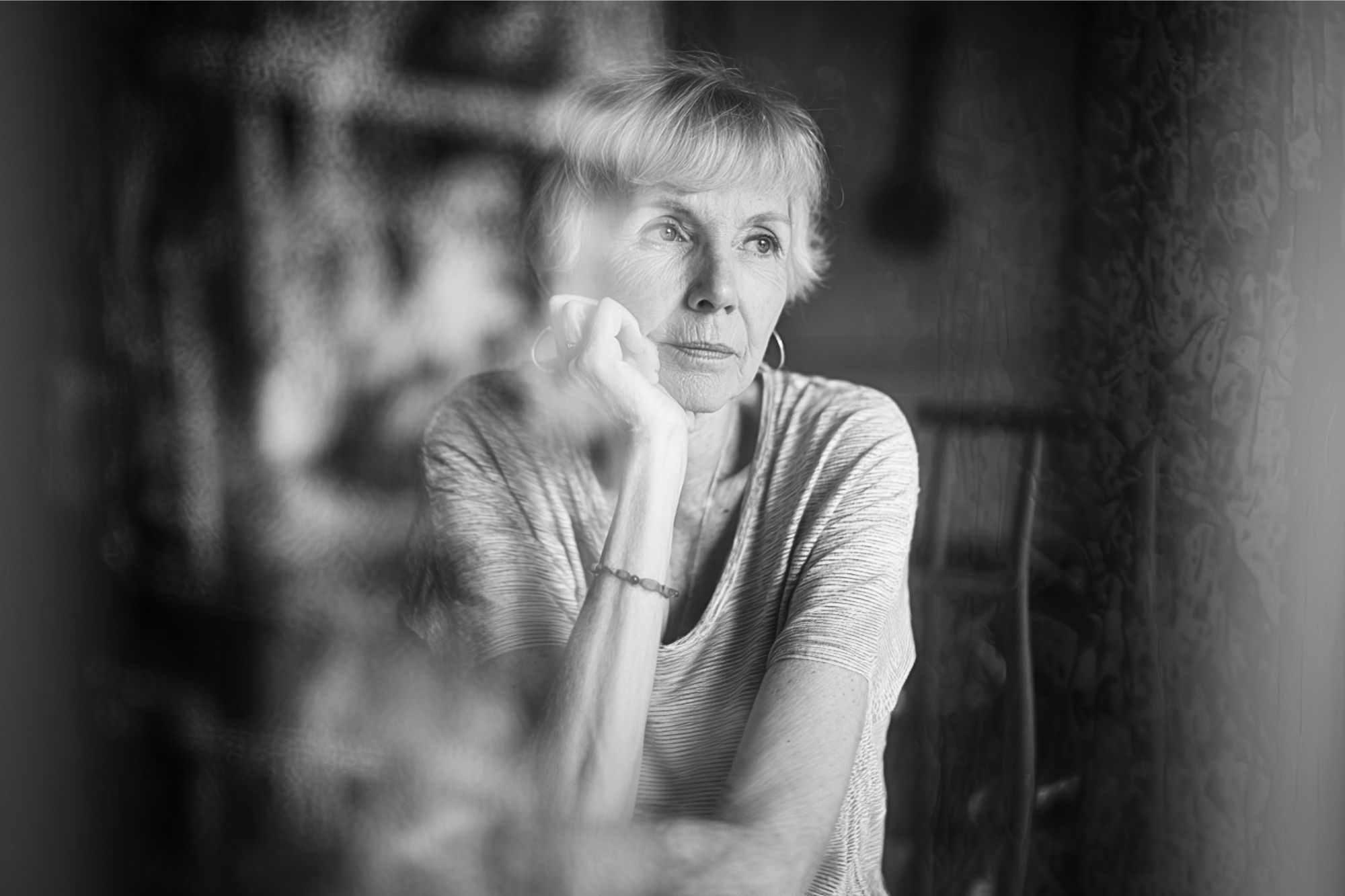 Black and white photo of an older woman with short hair, resting her chin on her hand and gazing thoughtfully out of a window. She appears pensive and lost in thought.