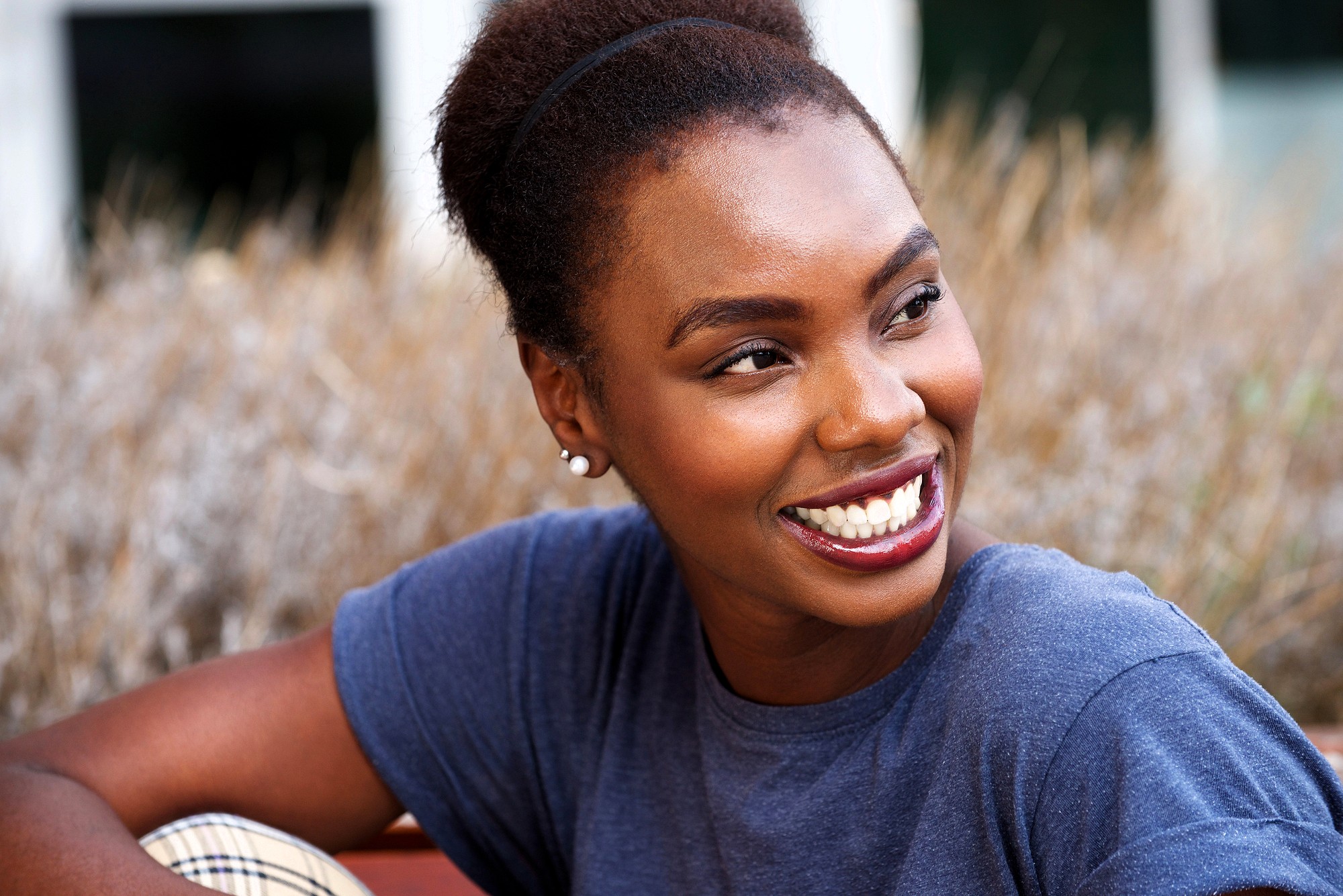 A young woman with dark skin and natural hair smiles brightly while looking to the side. She is wearing a blue shirt and stud earrings, sitting outdoors in front of blurred plants.