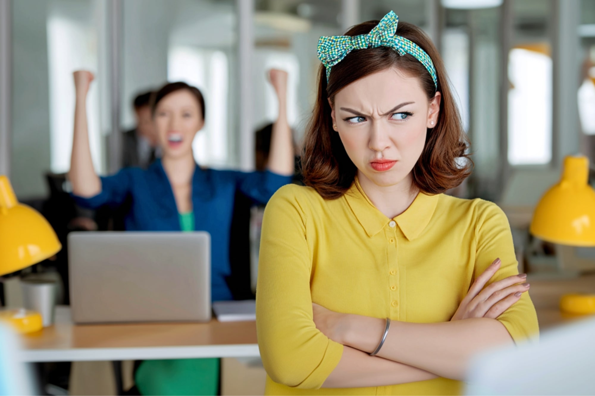 A woman in a yellow shirt stands with arms crossed, looking annoyed, while another woman in the background celebrates with raised fists at a desk in an office setting.