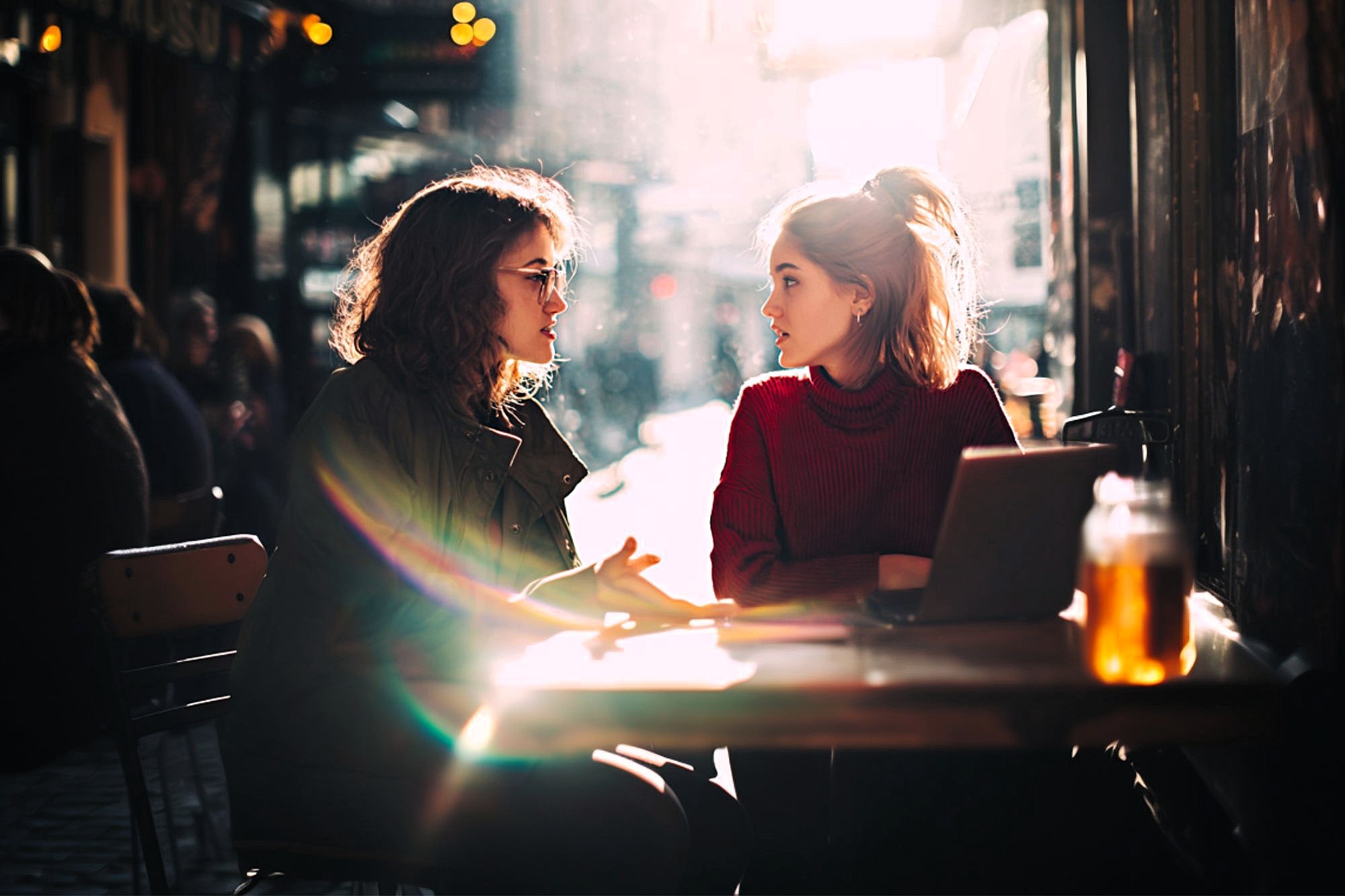 Two women sit at an outdoor café table, talking. One gestures while the other listens. Sunlight streams in, creating lens flare. A laptop and drink jar are on the table. The scene feels warm and casual.