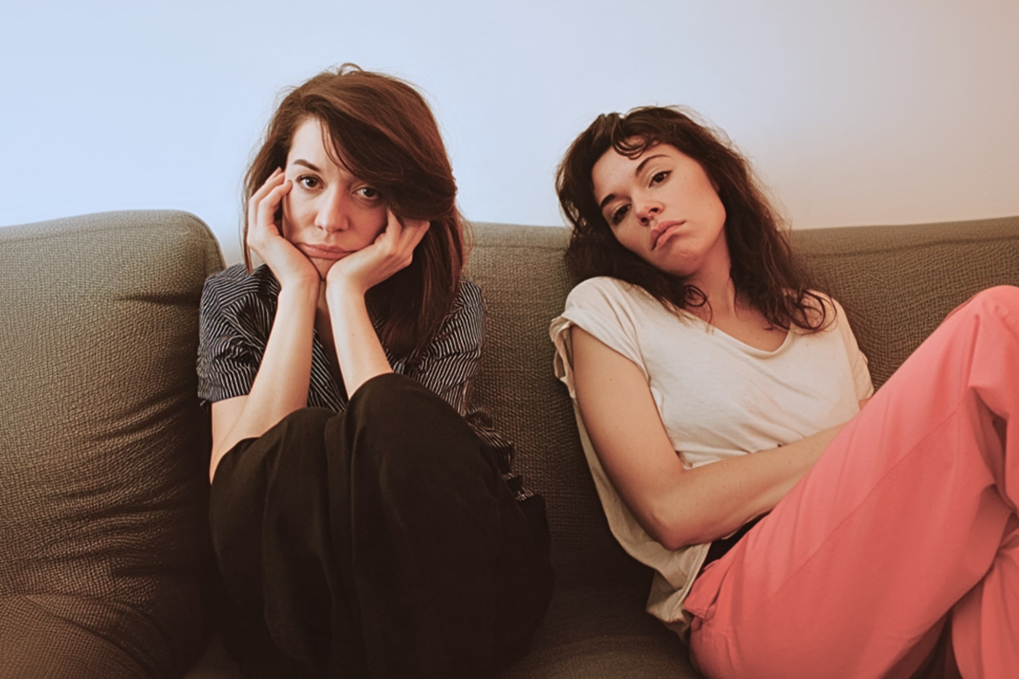 Two women sit on a couch indoors. One woman, with dark hair and striped shirt, rests her face in her hands and looks at the camera. The other, in a white t-shirt and pink pants, leans back with a neutral expression.