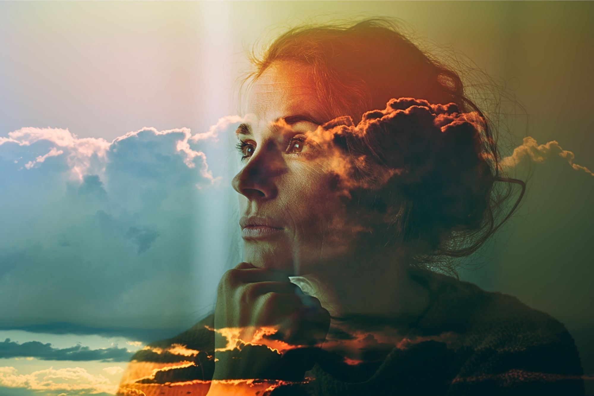 A double exposure image of a woman gazing thoughtfully into the distance, overlaid with colorful clouds and sunlight, creating a dreamy, contemplative atmosphere.