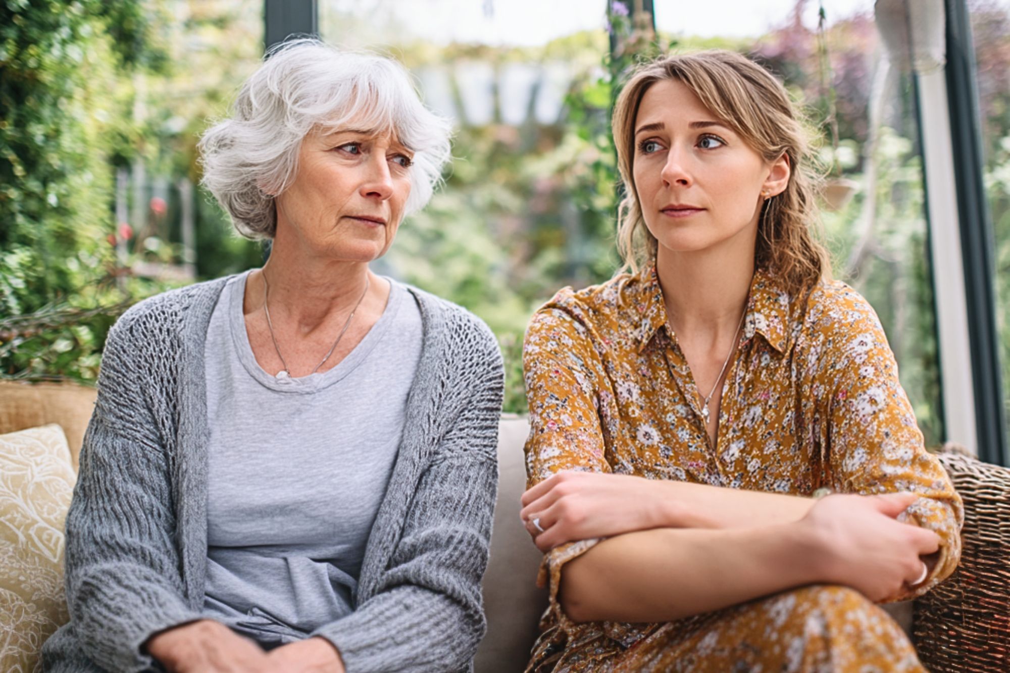 An older woman with gray hair and a younger woman with light brown hair sit next to each other indoors, both looking concerned and thoughtful. The younger woman has her arms crossed, and greenery is visible outside the windows.
