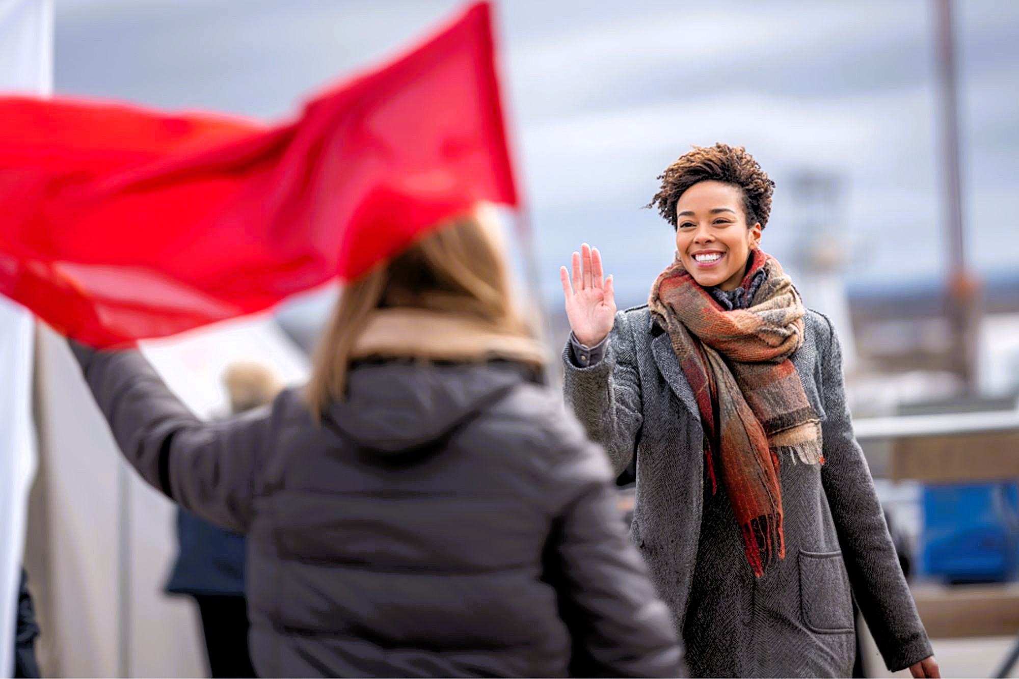 A smiling woman in a coat and scarf waves at someone holding a red flag outdoors on a cloudy day.